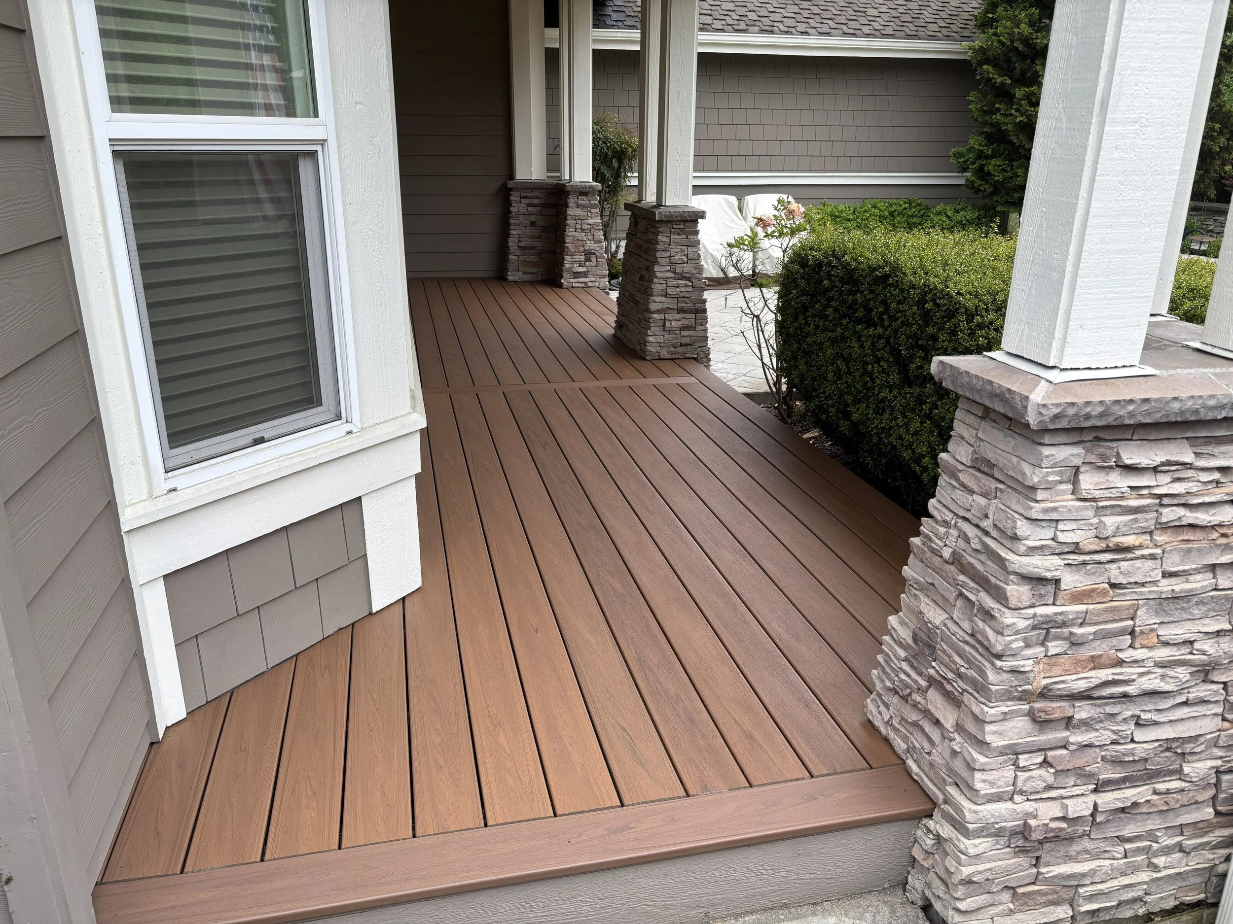 Close-up view of a wooden porch with stone pillars, a window on the left, and bushes on the right.
