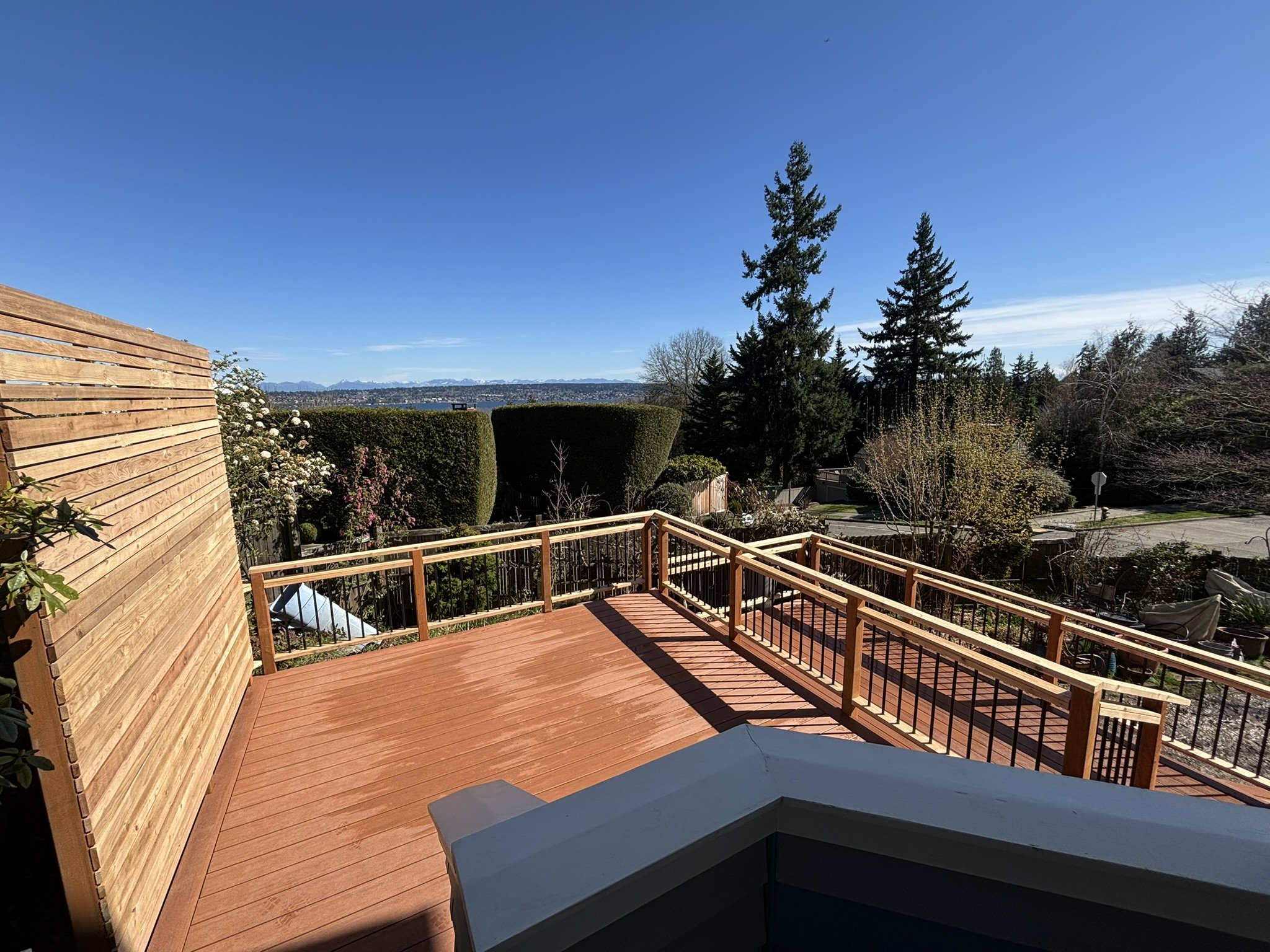 View of a wooden deck with railing, overlooking a suburban neighborhood with trees and a distant cityscape under a blue sky.