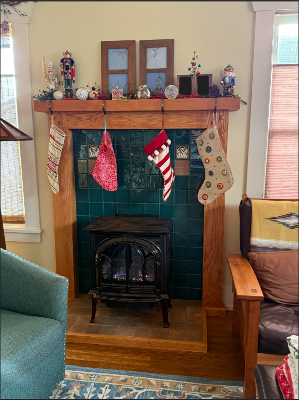 Christmas decorations on a wooden fireplace mantel with a small electric fireplace below. Four Christmas stockings hang from the mantel, and holiday figurines, ornaments, and a small decorated tree adorn the top. The room has yellow walls, a window with a blind on the right, and a blue armchair partially visible on the left.