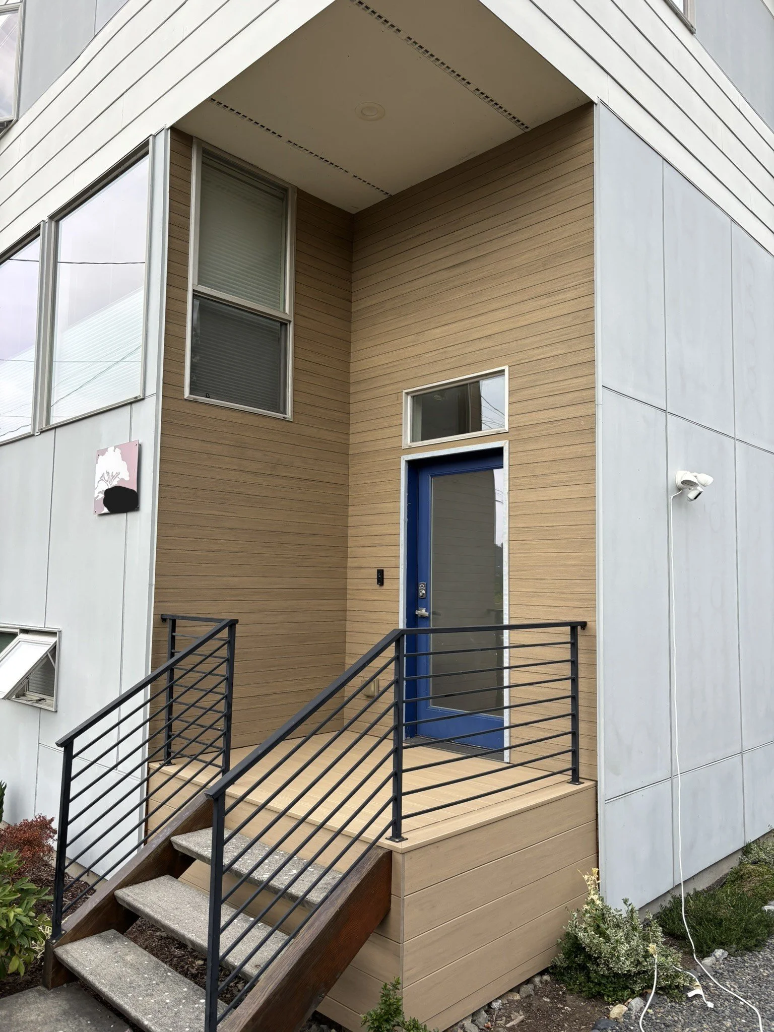 Modern apartment building entrance with a small staircase, black metal railing, blue door, and beige and gray exterior walls.