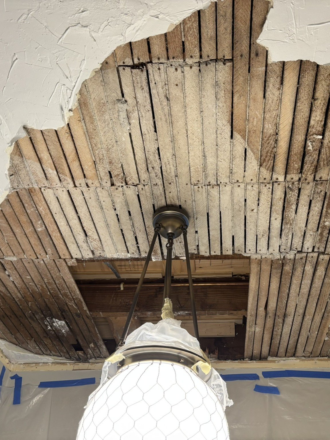 Ceiling renovation work showing a partially removed ceiling with exposed wooden beams and bricks, with a chandelier hanging from it.