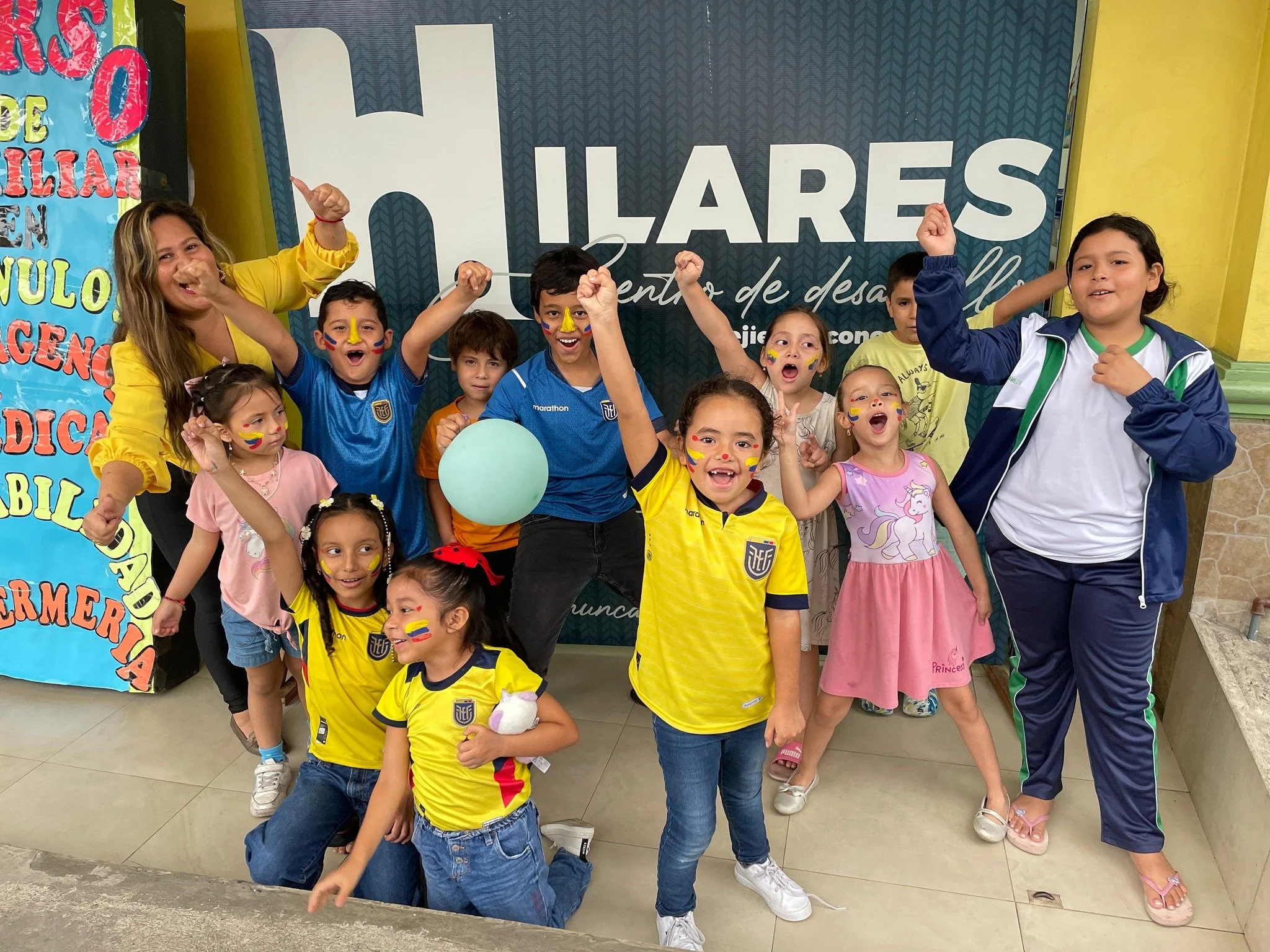 Grupo de niños y una mujer con alegría, vestida con camisetas amarillas y ropa de deportes, celebrando con los brazos en alto en un evento escolar, algunos con cara pintada y globos, en un fondo con letreros coloridos.