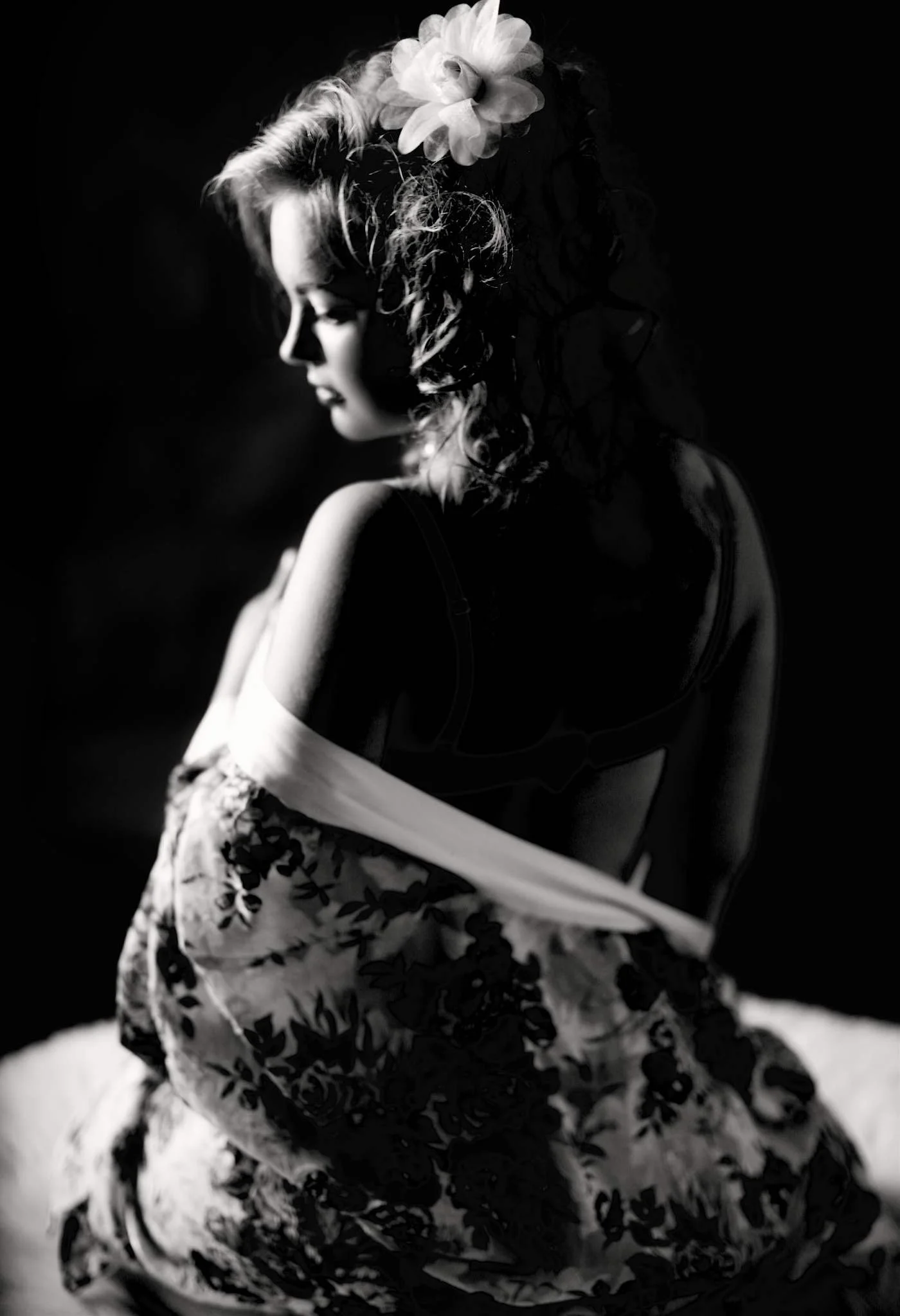A black and white photograph of a woman with curly hair, wearing a large flower headpiece, a dark top with thin straps, and a floral-patterned skirt, sitting with her arms crossed and looking down.