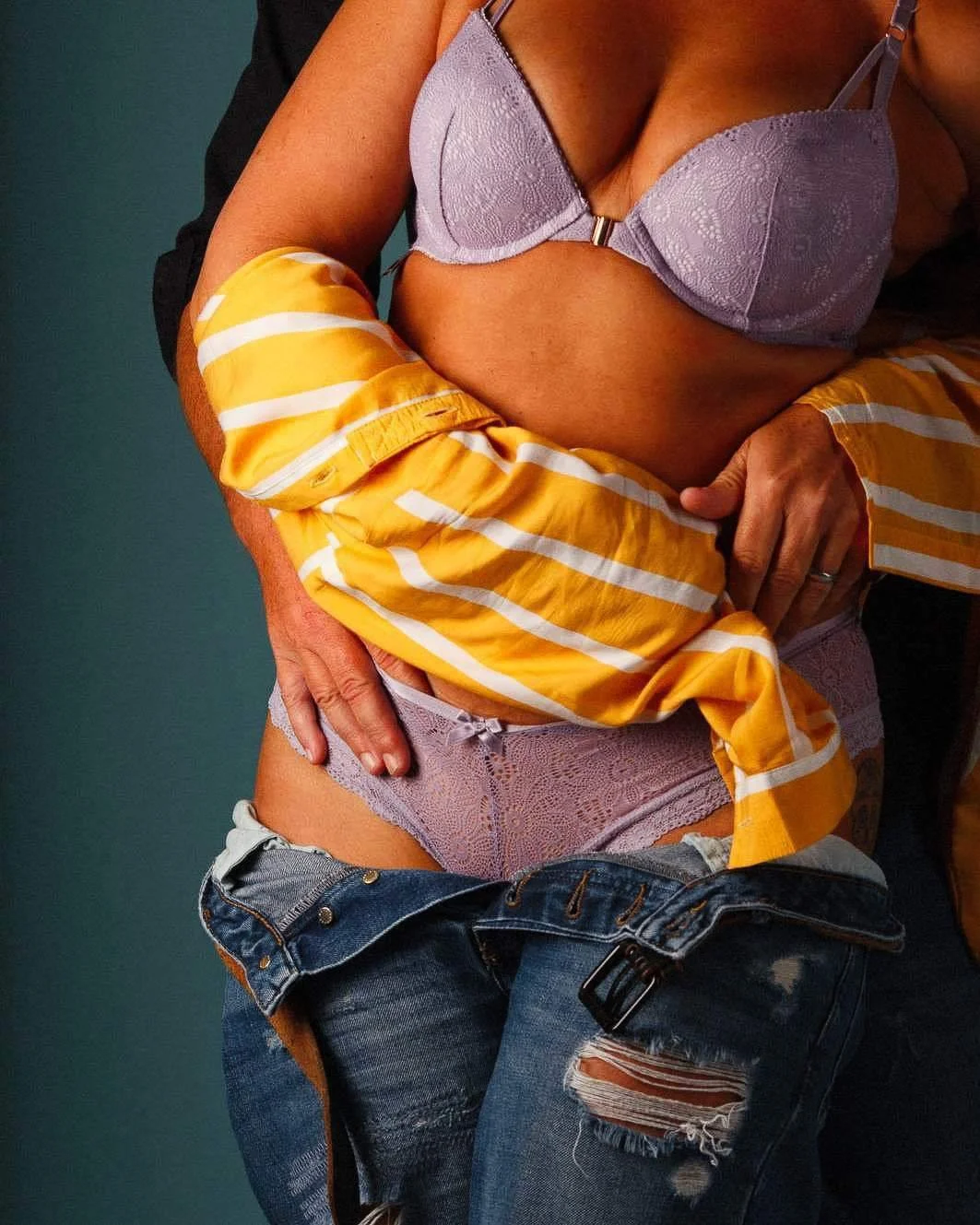 Close-up of a woman wearing purple lace lingerie and ripped jeans, being embraced by a person with wearing a yellow striped shirt, against a teal background.
