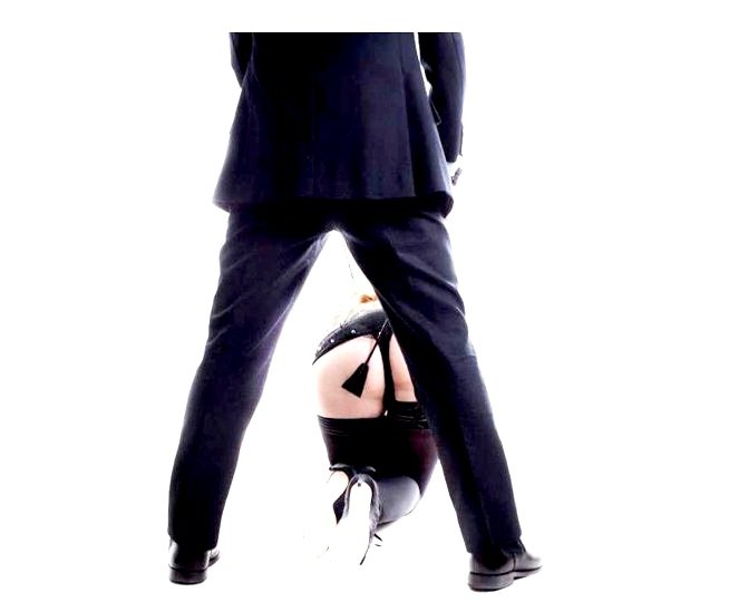 A person in a suit standing over another person who is kneeling, wearing black thigh-high stockings and black high heels.