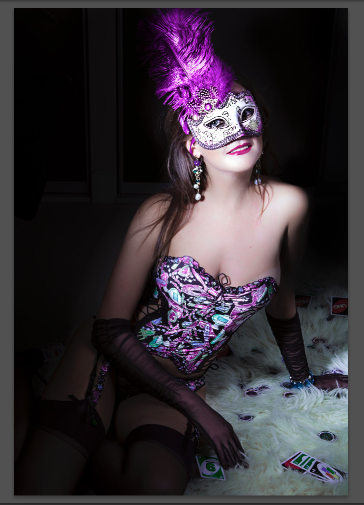 Woman wearing a colorful, patterned corset, sheer black gloves, and black stockings, sitting on a fluffy surface with scattered UNO cards, wearing a purple feathered masquerade mask with rhinestones and earrings with blue and white beads, smiling at 
