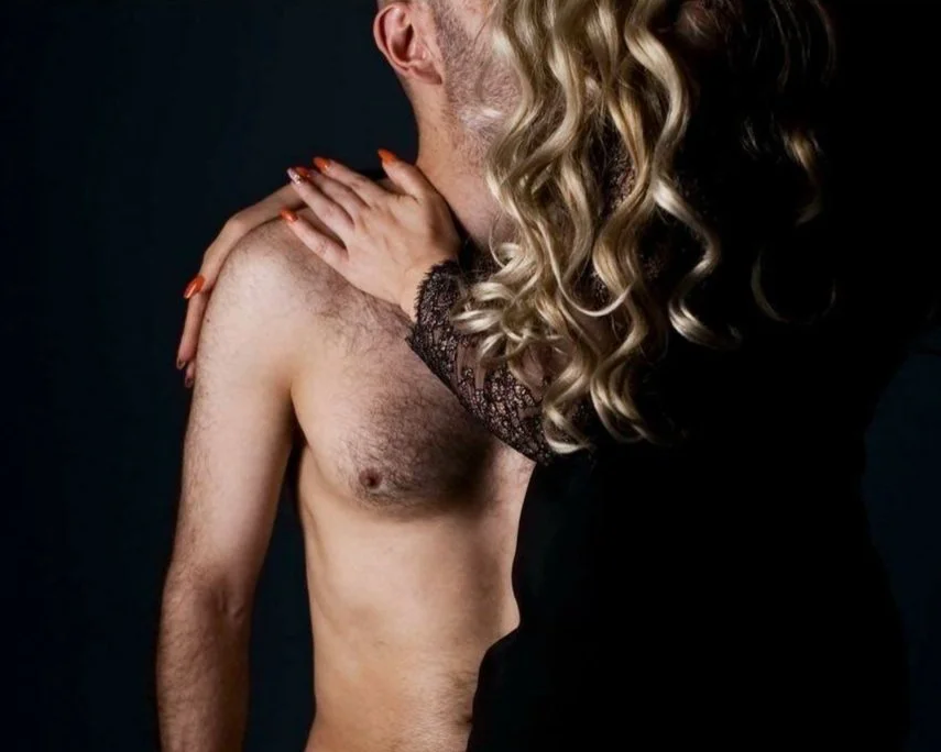 Close-up of a shirtless man with a woman leaning in, her hand on his shoulder, against a dark background.