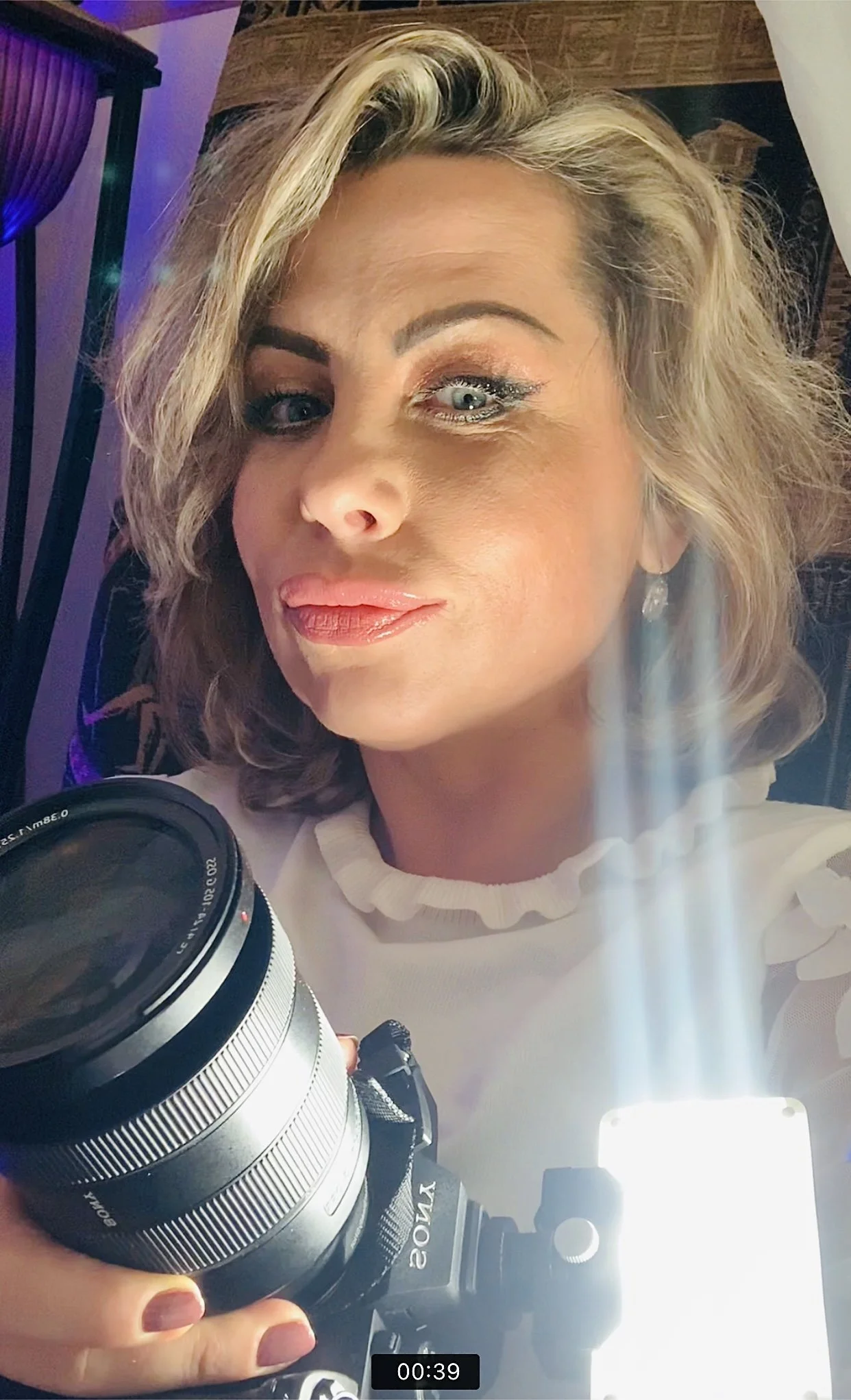 Close-up of a person with wavy blonde hair holding a camera with a bright light attachment.