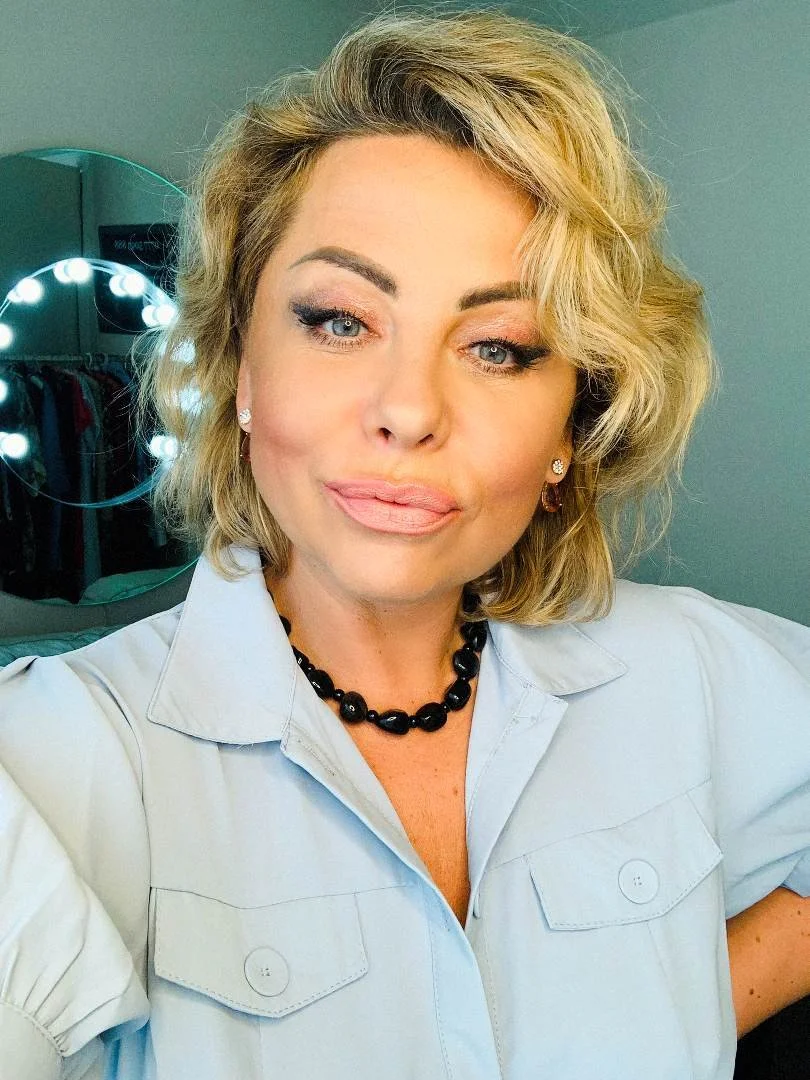 A woman with blonde wavy hair is taking a selfie in front of a mirror with vanity lights. She is wearing a light-colored shirt, a black bead necklace, and earrings.