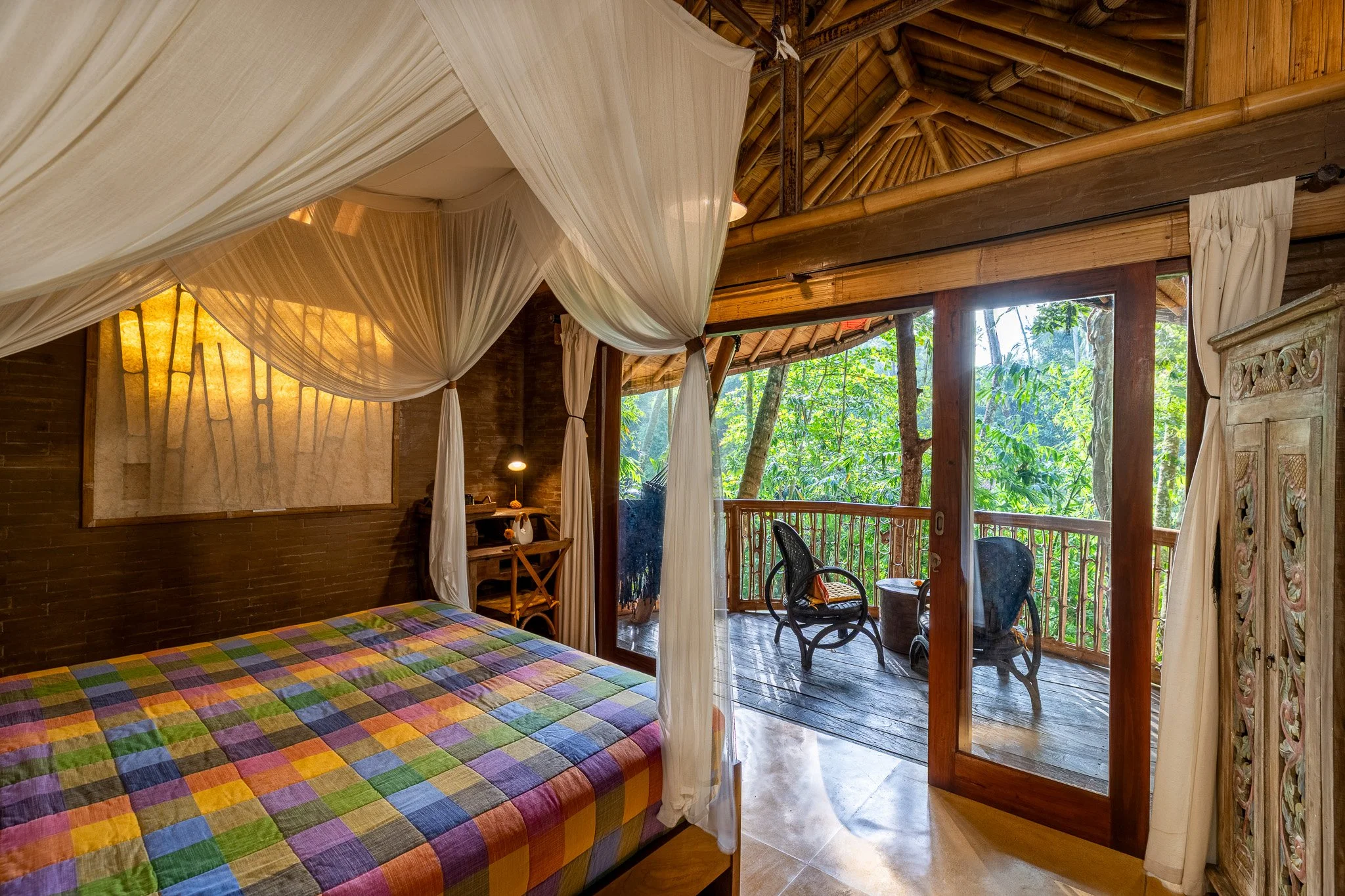 Interior of a rustic bedroom with a colorful patchwork bedspread, mosquito net canopy, and a wooden balcony with outdoor chairs overlooking lush green trees.