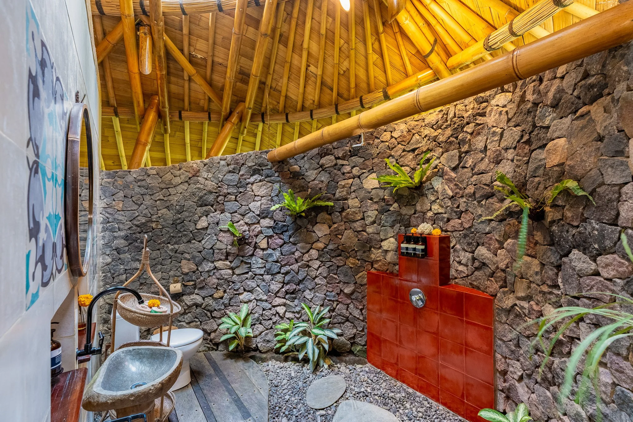 A rustic outdoor bathroom with a stone wall, bamboo ceiling, and plants; includes a small sink, a toilet with flowers, and a shower area.