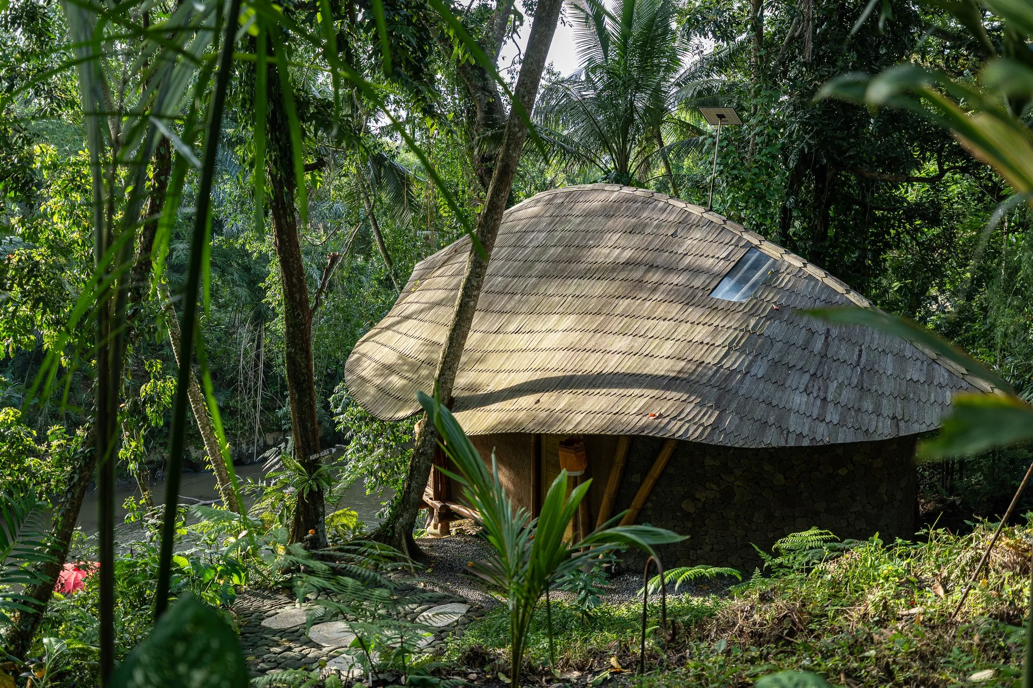 A unique round hut with a shingled roof is situated among lush green tropical trees and plants in a forest.