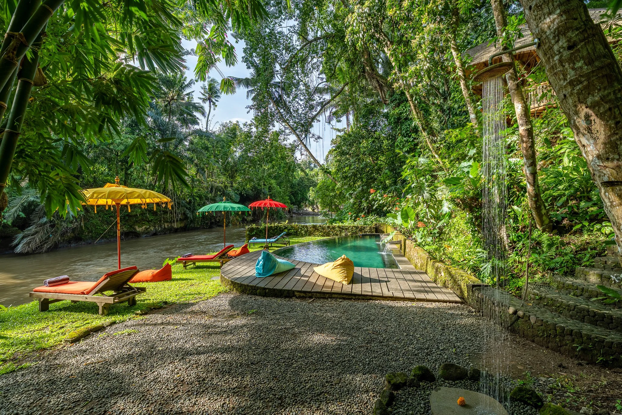 A tropical outdoor pool area with a wooden deck, several lounge chairs, colorful umbrellas, beans bags, lush green foliage, and a waterfall feature.