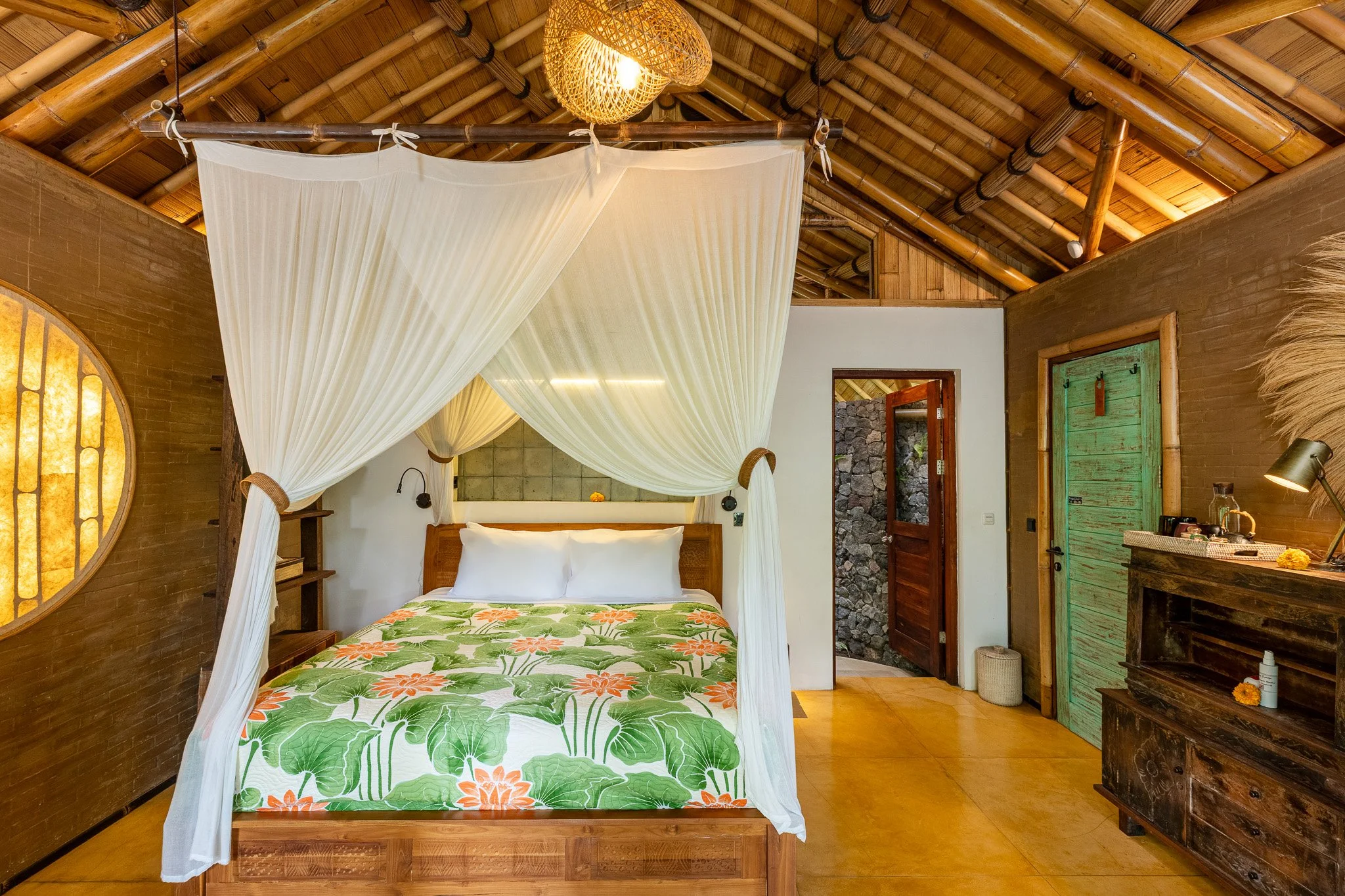 A bedroom with a four-poster bed draped with white curtains, a green and orange floral bedspread, wooden furniture, a green door, and a rustic ceiling with bamboo beams.