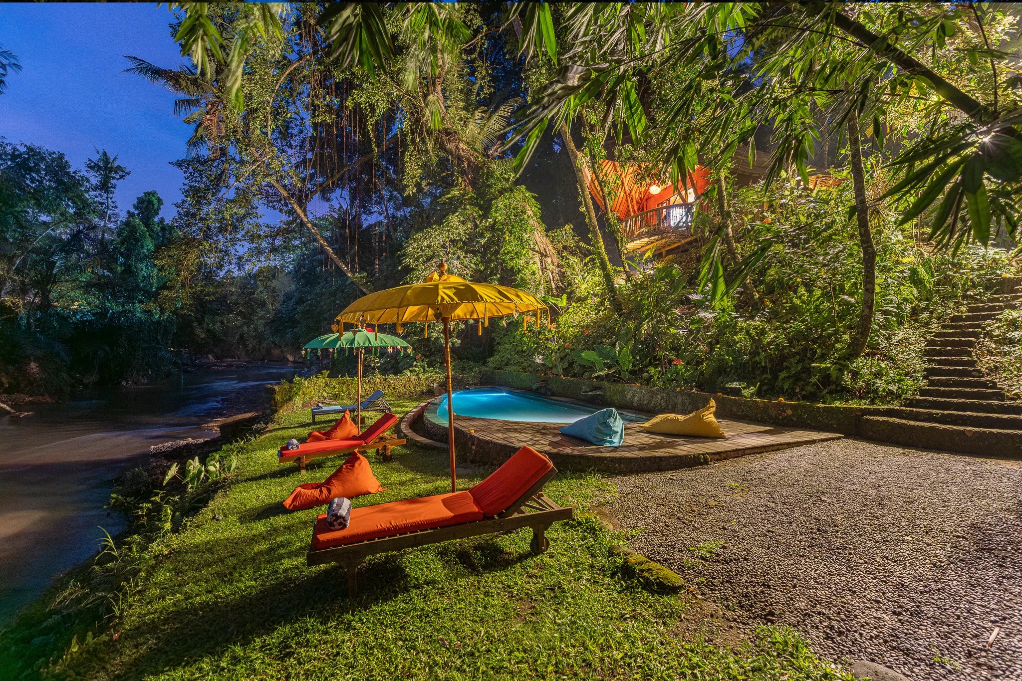 Nighttime view of a tropical outdoor pool area surrounded by lush greenery, with outdoor lounge chairs, umbrellas, colorful bean bags, and a small staircase leading into the woods.
