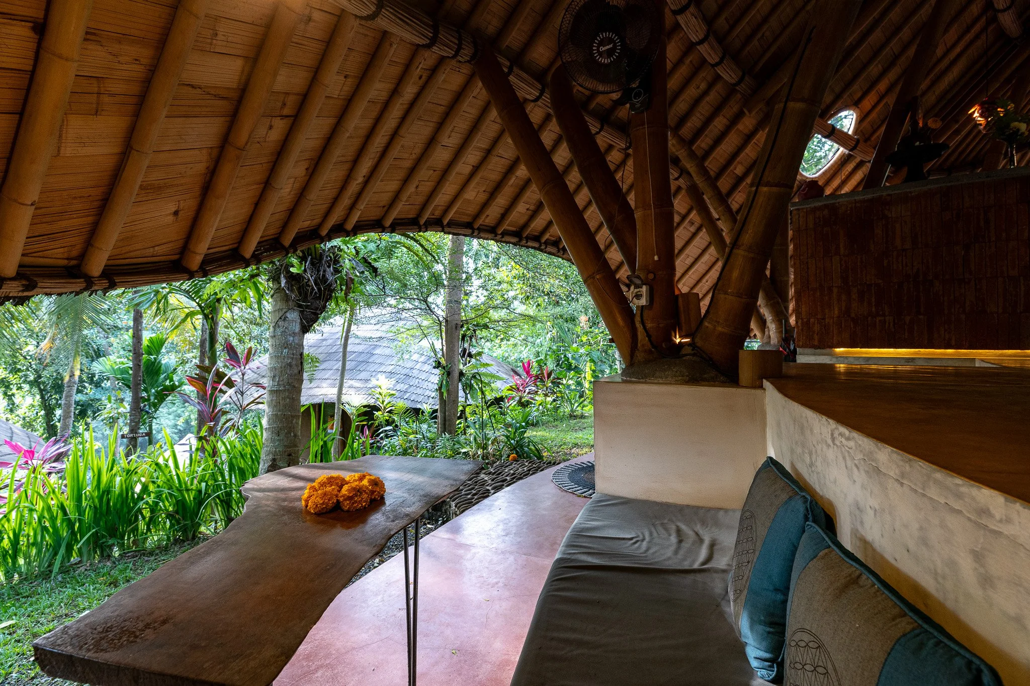 Interior of a bamboo and wood structure overlooking a lush garden with green plants and trees, a table with orange flowers, and seating area.