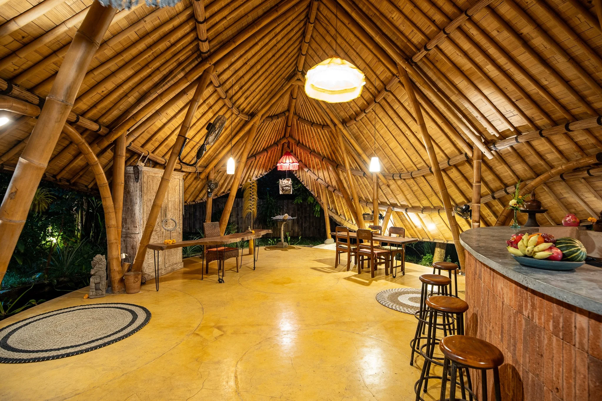 Interior of a bamboo hut with warm lighting, wooden furniture, and tropical decor, including a bar counter with a fruit bowl and sitting stools.