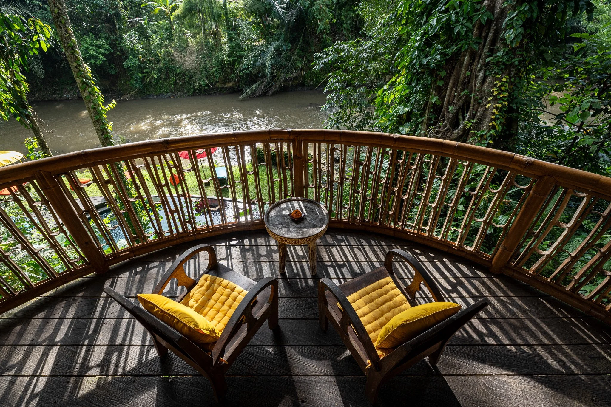 A wooden balcony overlooking a river surrounded by lush green tropical trees. Two chairs with yellow cushions and a small round table with a tray are on the balcony, with sunlight casting shadows on the floor.