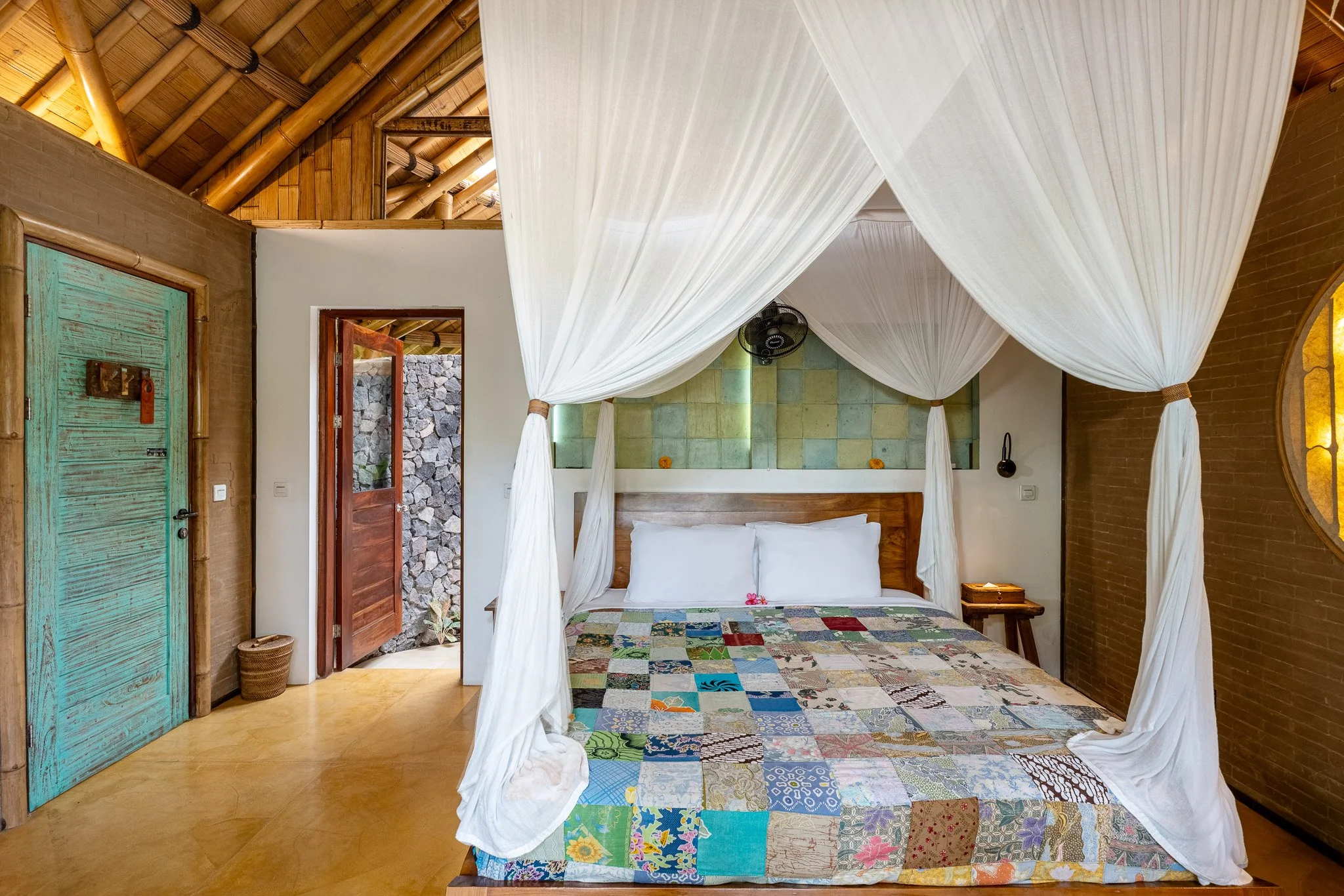 A bedroom with a four-poster bed draped with white curtains, a patchwork quilt, a wooden headboard, a small nightstand with a lamp, a textured green wall behind the bed, and an open door leading outside with stone walls visible.