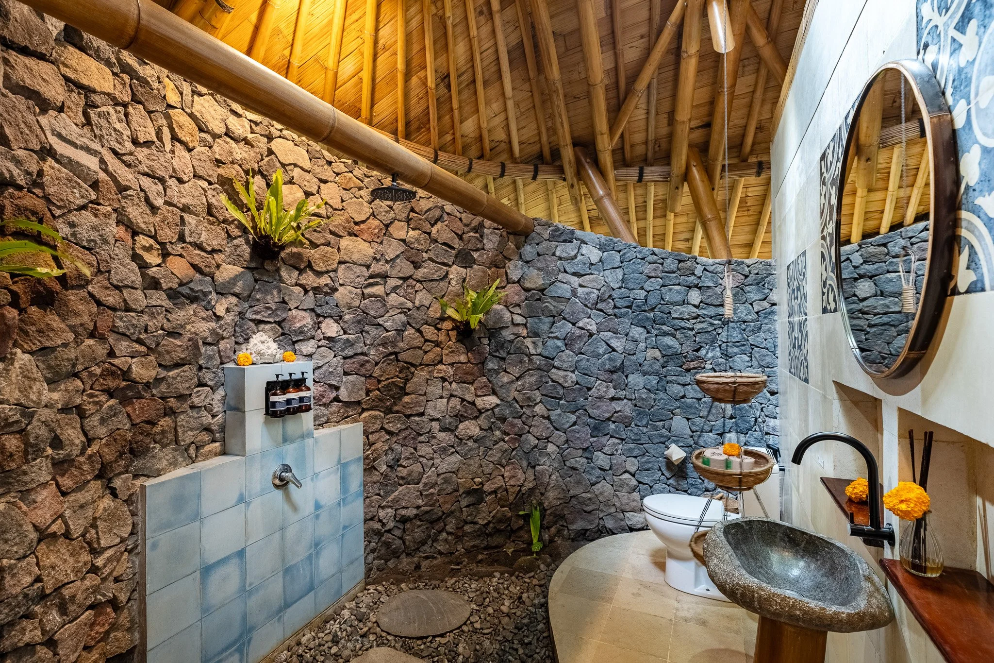 Bamboo and stone bathroom with a rustic sink, plants, and a shower area with rocks and a showerhead.