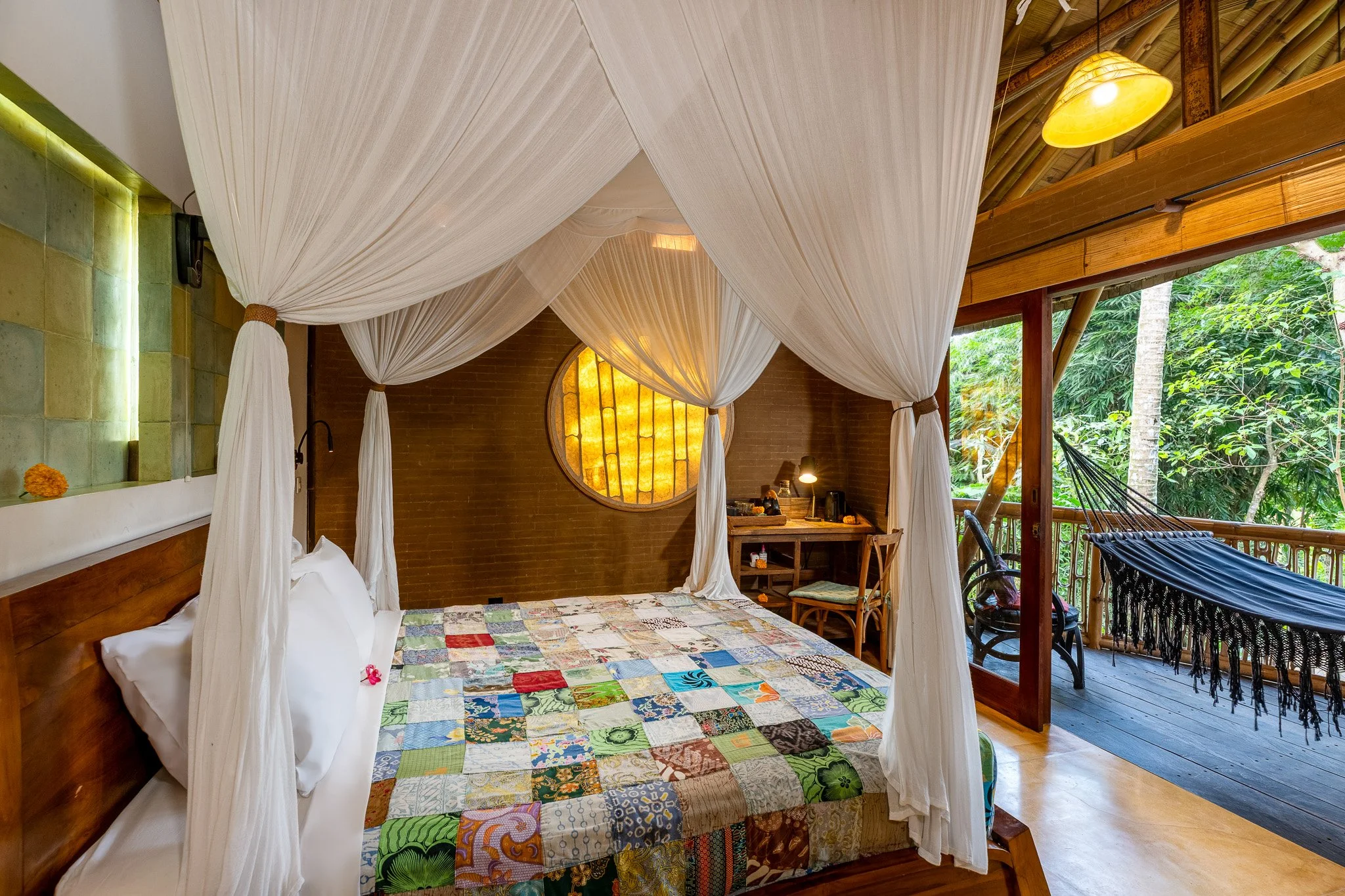 Bedroom with a four-poster bed covered in a colorful quilt, a small workspace with a desk and chair, and a balcony with a hammock and lush greenery outside.