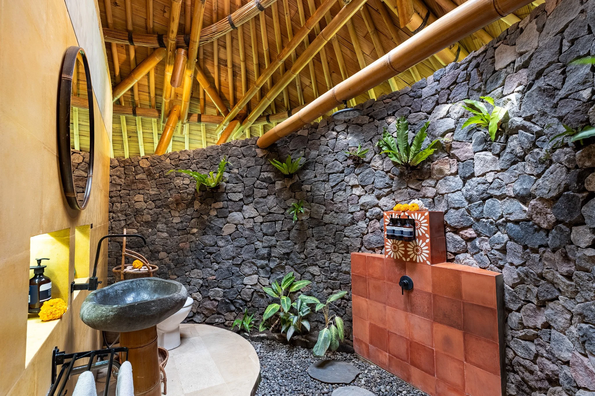 Outdoor bathroom with a stone wall, plants, bamboo ceiling, and natural decor.