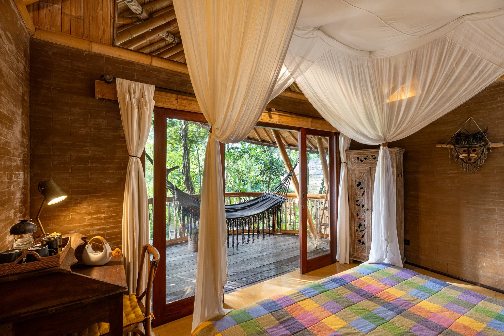 Room with tropical decor, featuring a bed with a colorful quilt, wooden furniture, and a balcony with a hammock surrounded by greenery.