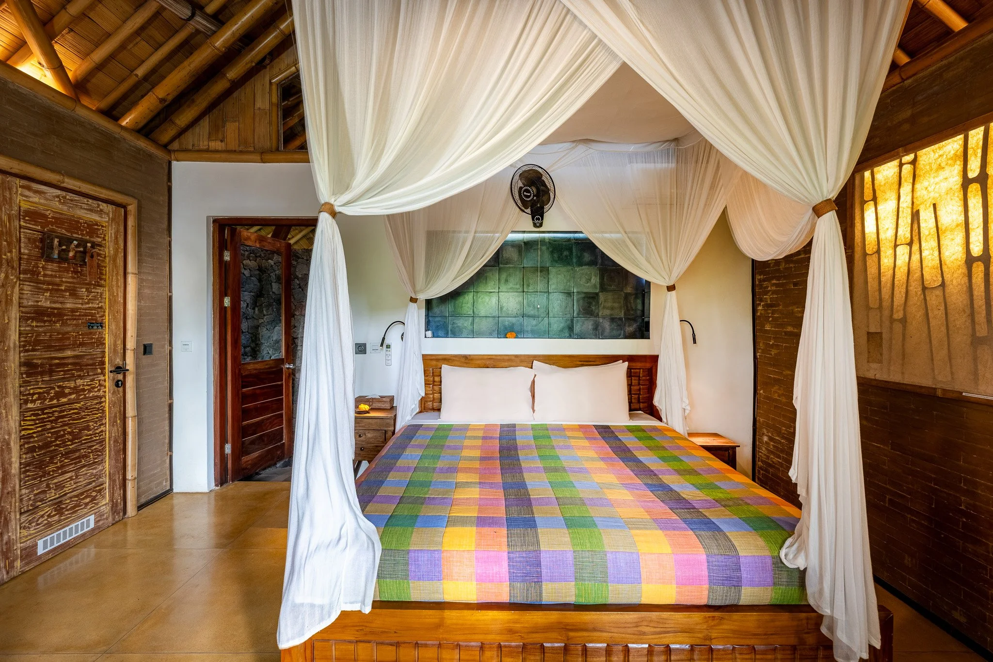 A bedroom with a four-poster bed covered in a colorful checkered bedspread, with white pillows. The room has a tropical vibe with a wooden door, rustic walls, and a thatched ceiling. Light filters through warm-colored wall panels on the right.