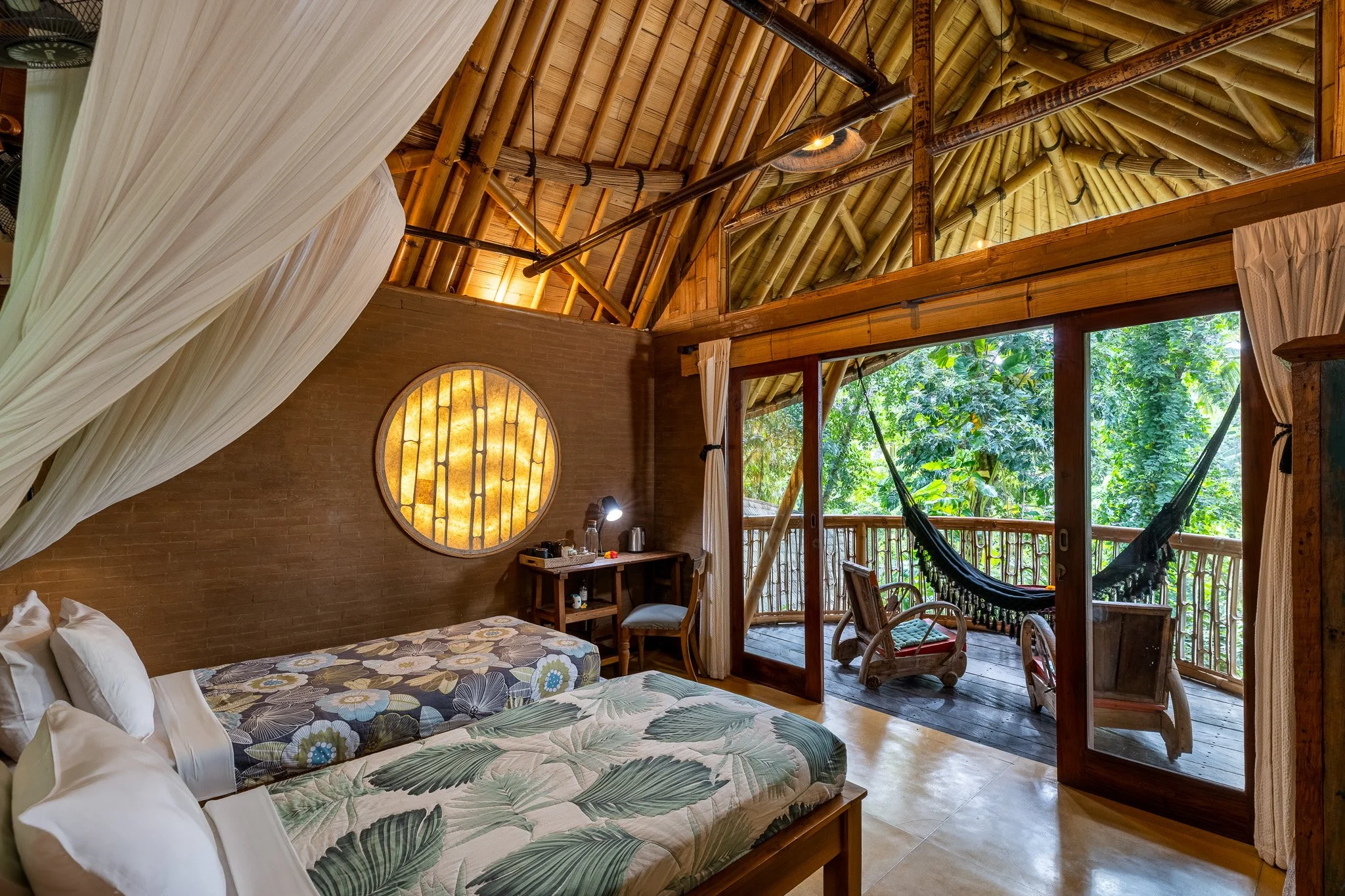 A cozy tropical bedroom with two beds, a small desk, and a balcony overlooking lush greenery. The room features a thatched ceiling, wooden furniture, and large sliding glass doors.