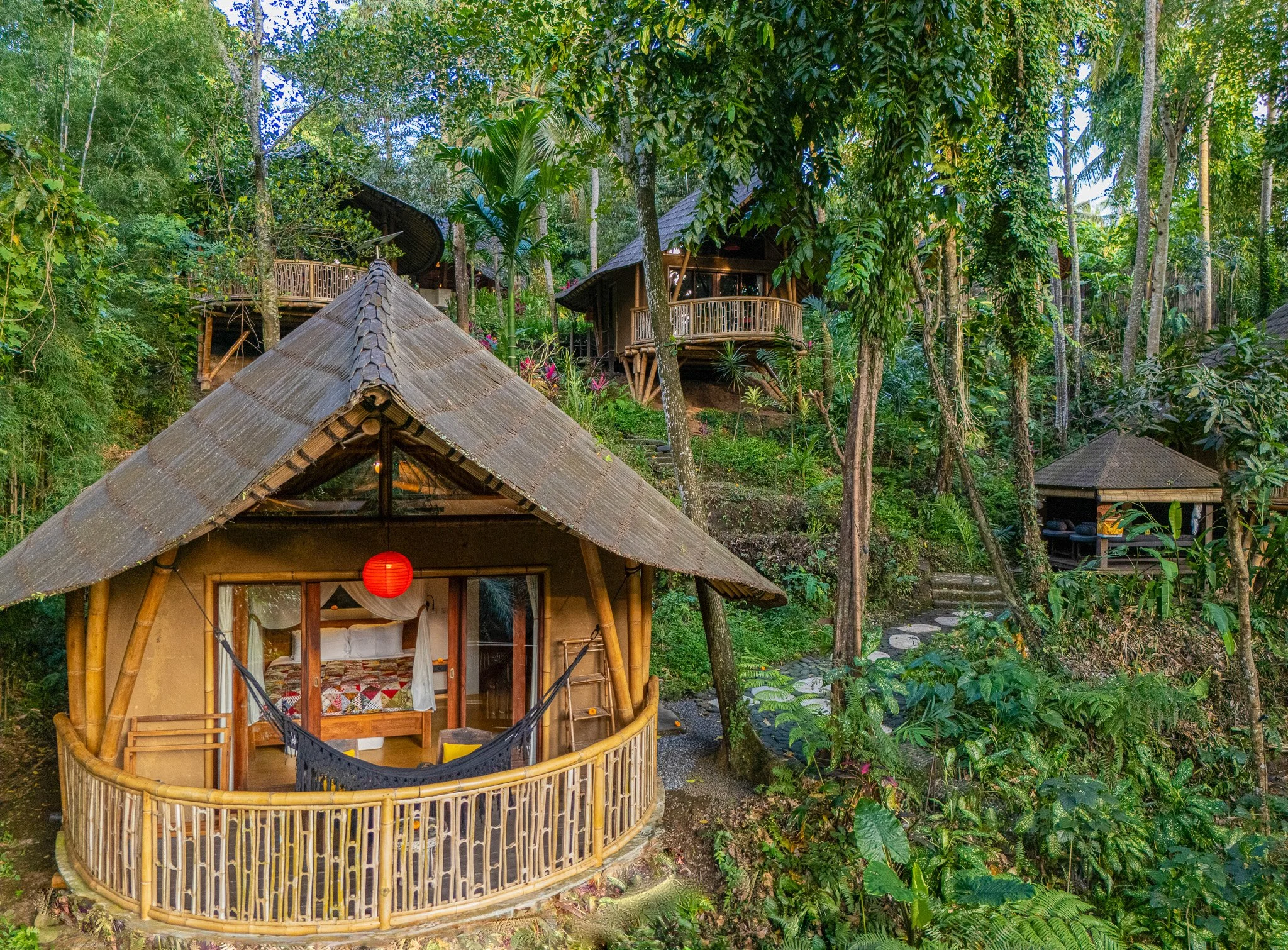 A bamboo and thatch-roof bungalow with a red paper lantern hanging outside, situated in a lush, green tropical forest with multiple tree houses and pathways in the background.
