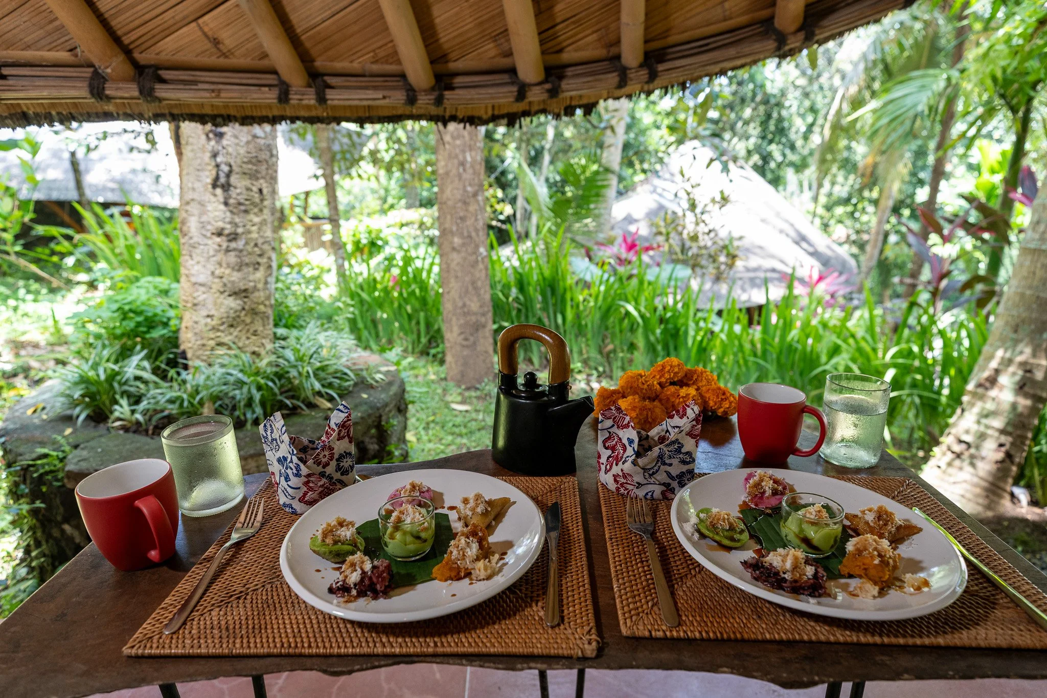 Two place settings with plates of food, cups, glasses, and a teapot on a wooden table outdoors with lush green plants and trees in the background.