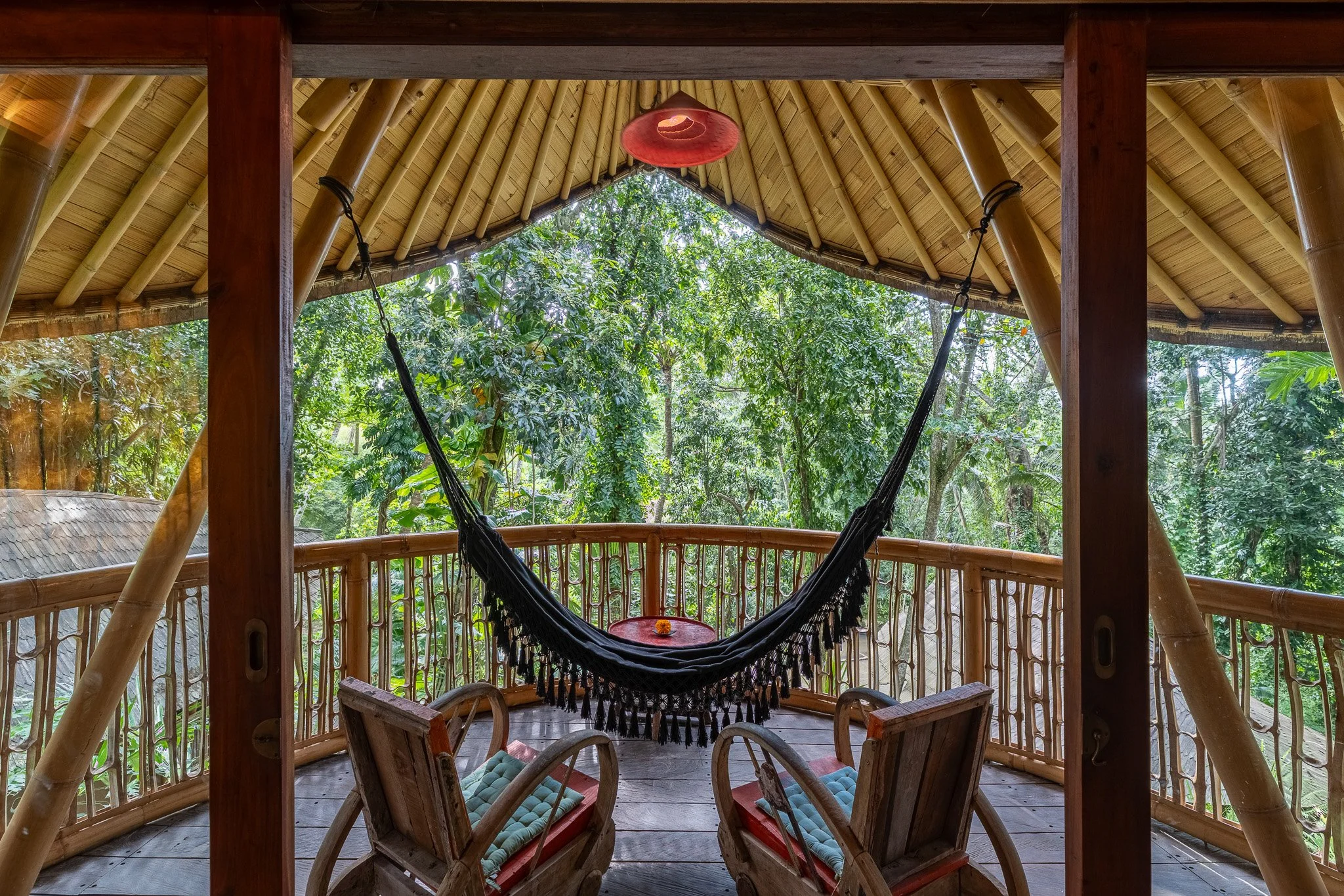 View of a bamboo balcony with a hammock, two wooden chairs, and a small round table, overlooking a lush green forest.