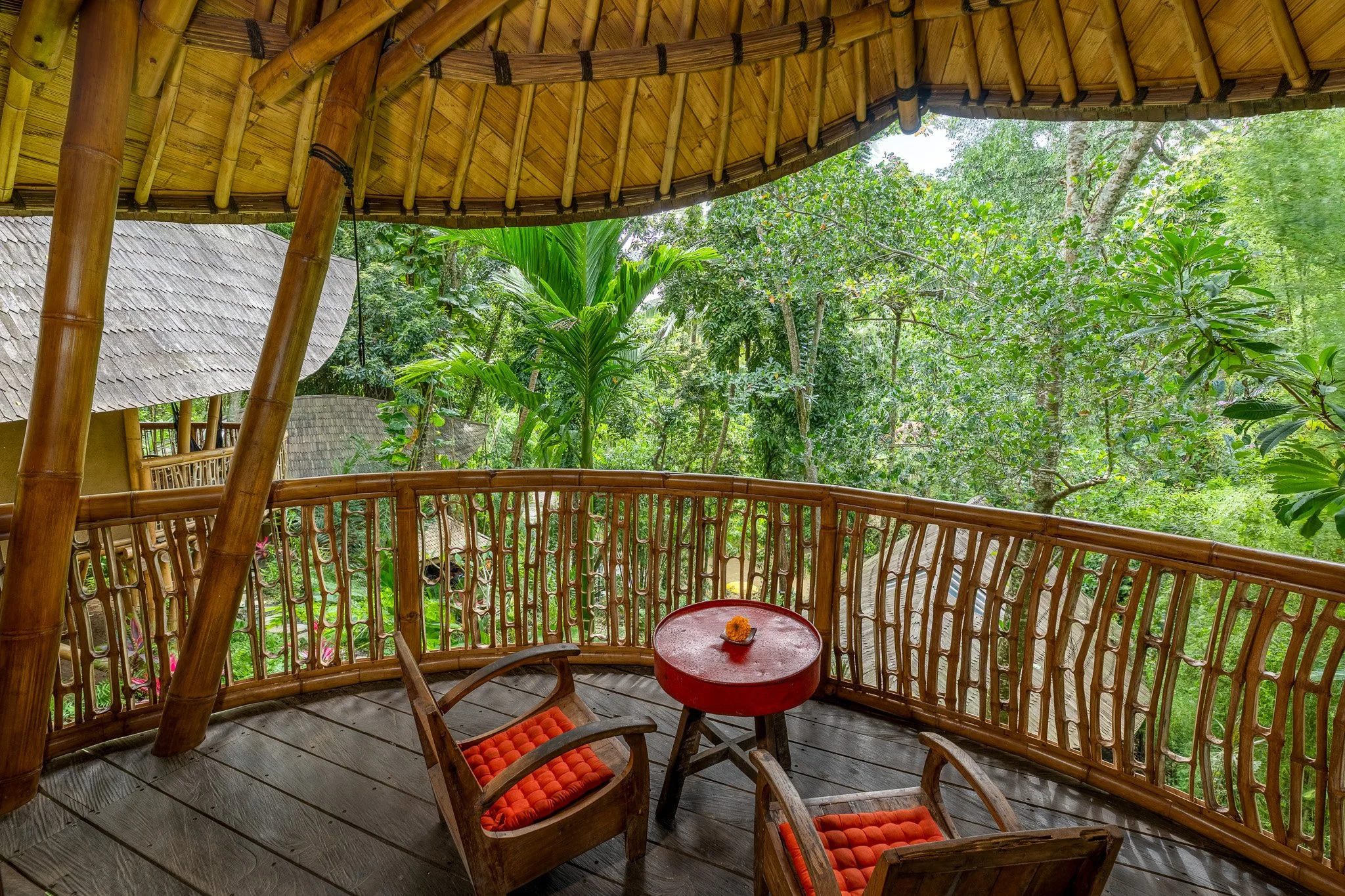 A bamboo balcony with a round red table and two wooden chairs with orange cushions, overlooking lush green tropical trees and plants.