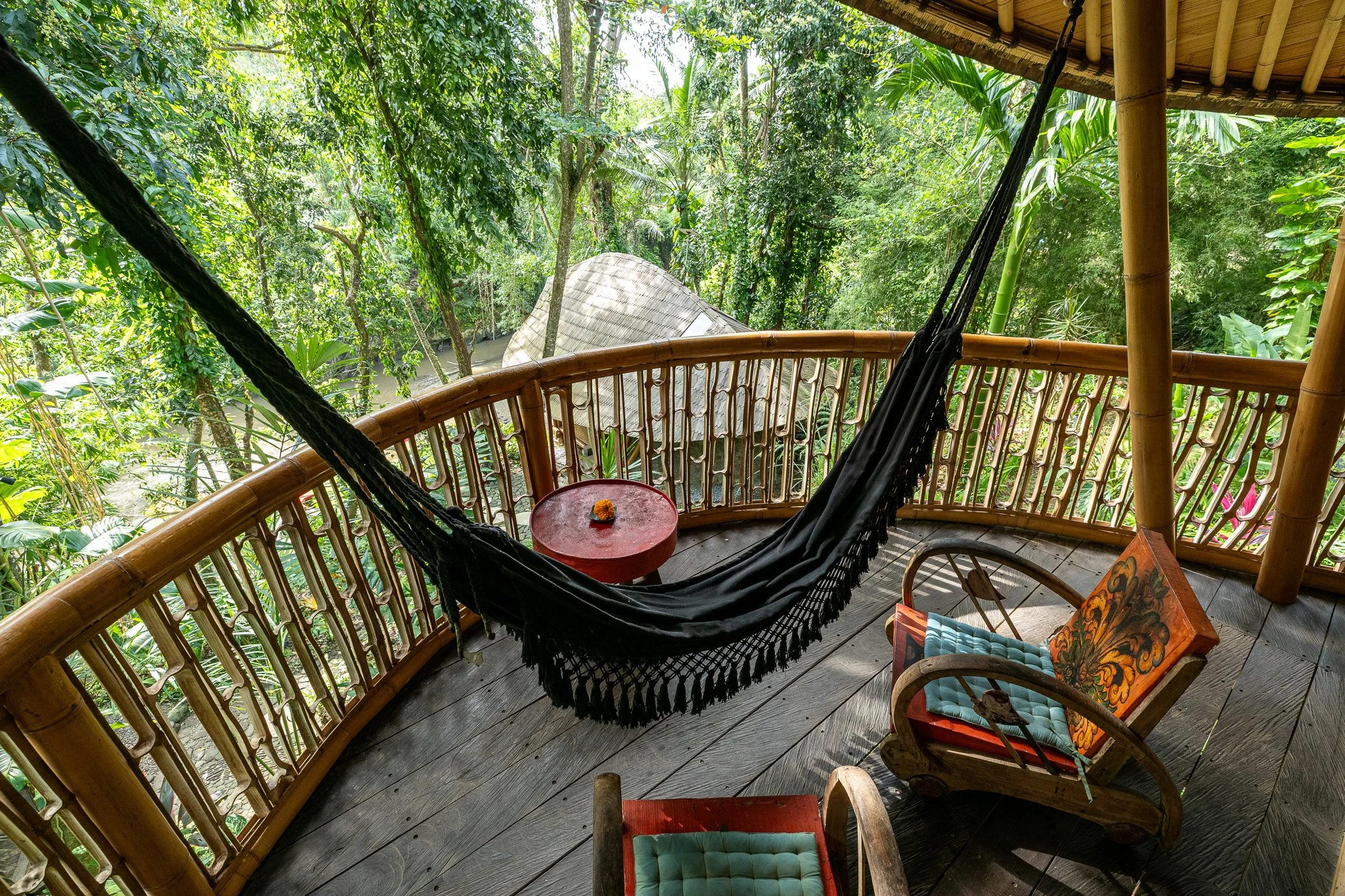 A balcony with a black hammock, two colorful wooden chairs with cushions, a small red round table, and surrounded by lush green jungle foliage.