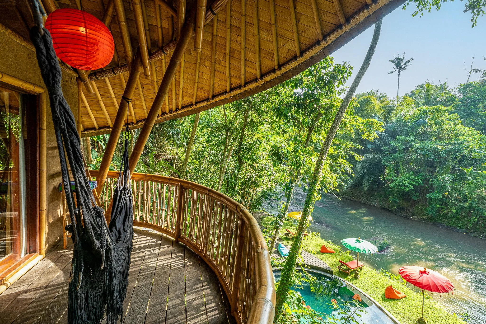 Bamboo balcony overlooking a river with lush green trees, colorful umbrellas, and lounge chairs
