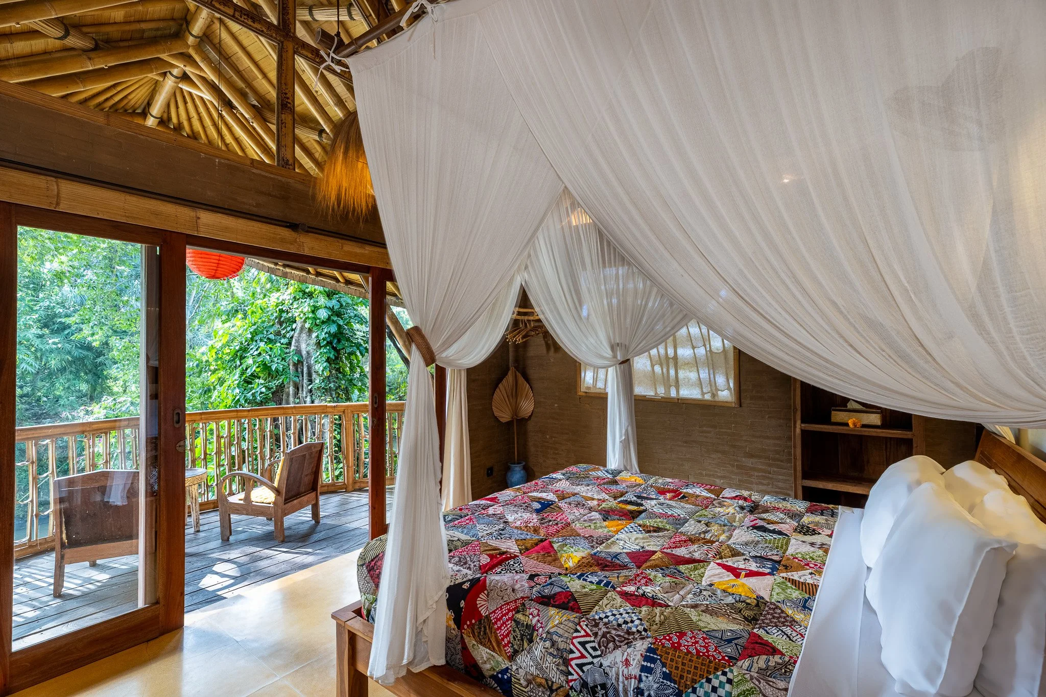 A bedroom with a four-poster bed decorated with a colorful patchwork quilt and white pillows, featuring a mosquito net canopy, and a balcony with outdoor seating, surrounded by lush greenery.