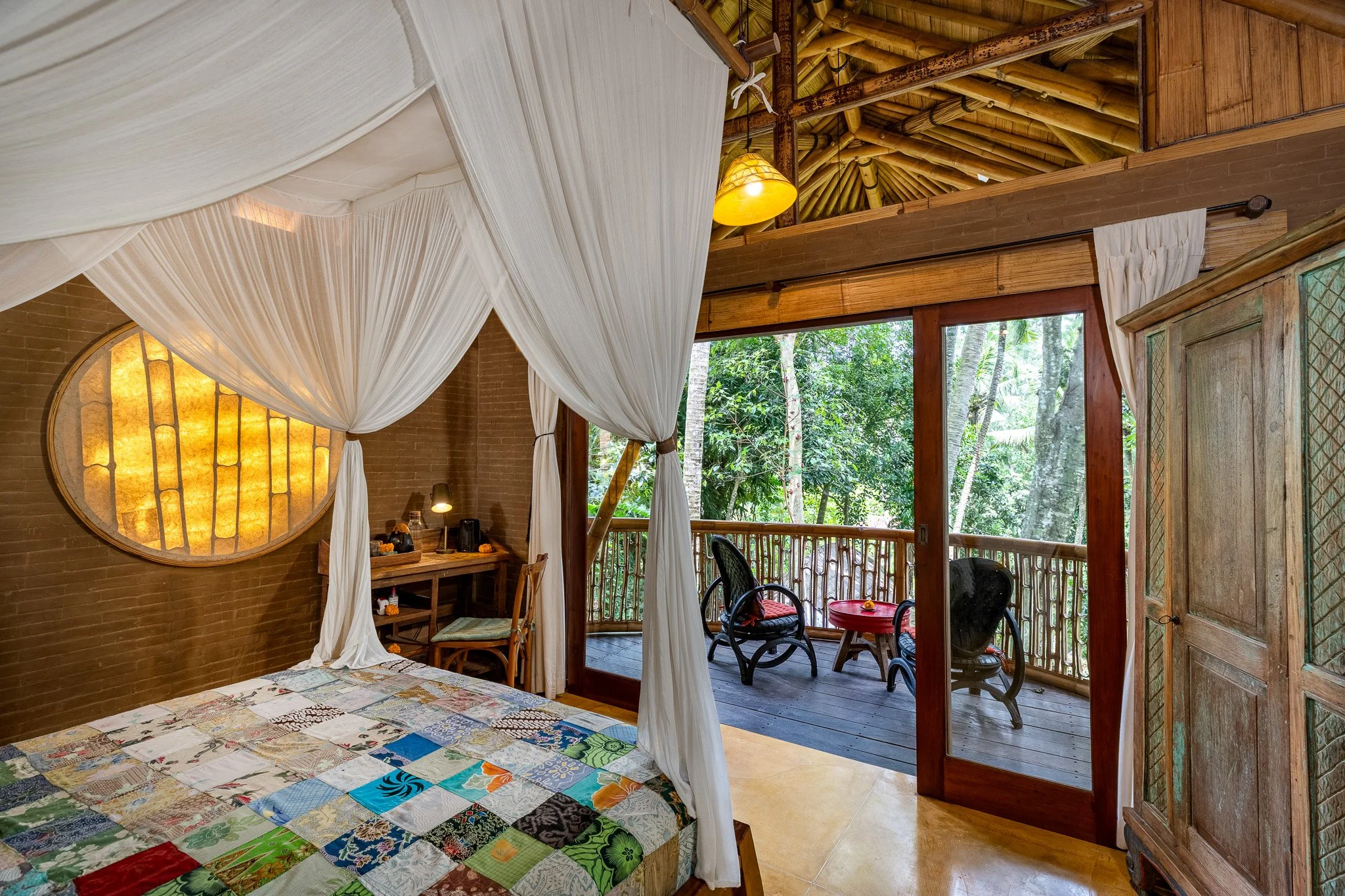 A cozy bedroom with a four-poster bed draped in white curtains, a wooden desk with a chair, and a balcony with outdoor seating surrounded by lush green trees.