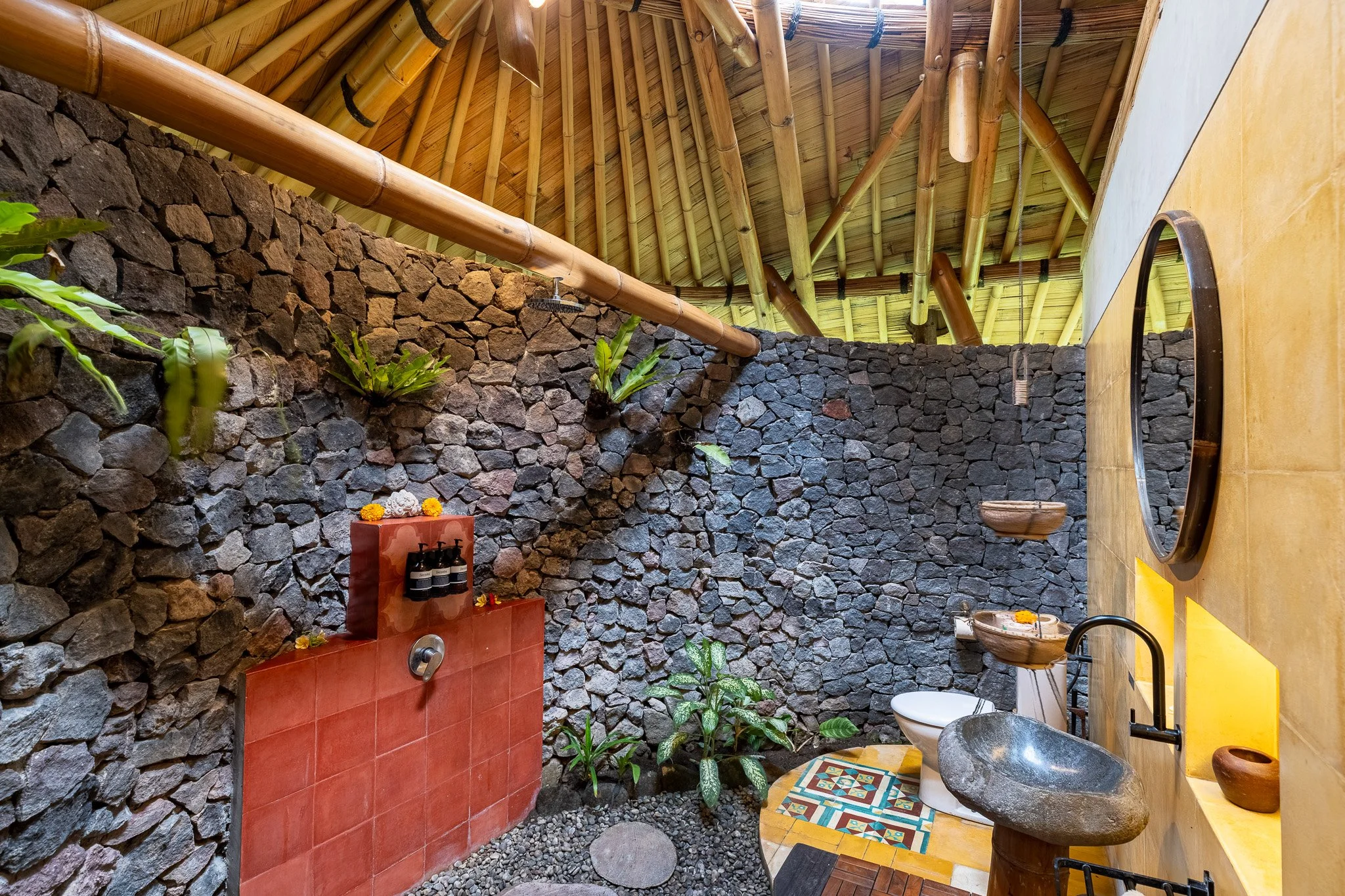 Outdoor bathroom with stone wall, bamboo roof, and plants. Features a stone sink, mirror, toilet, and decorative tiles.