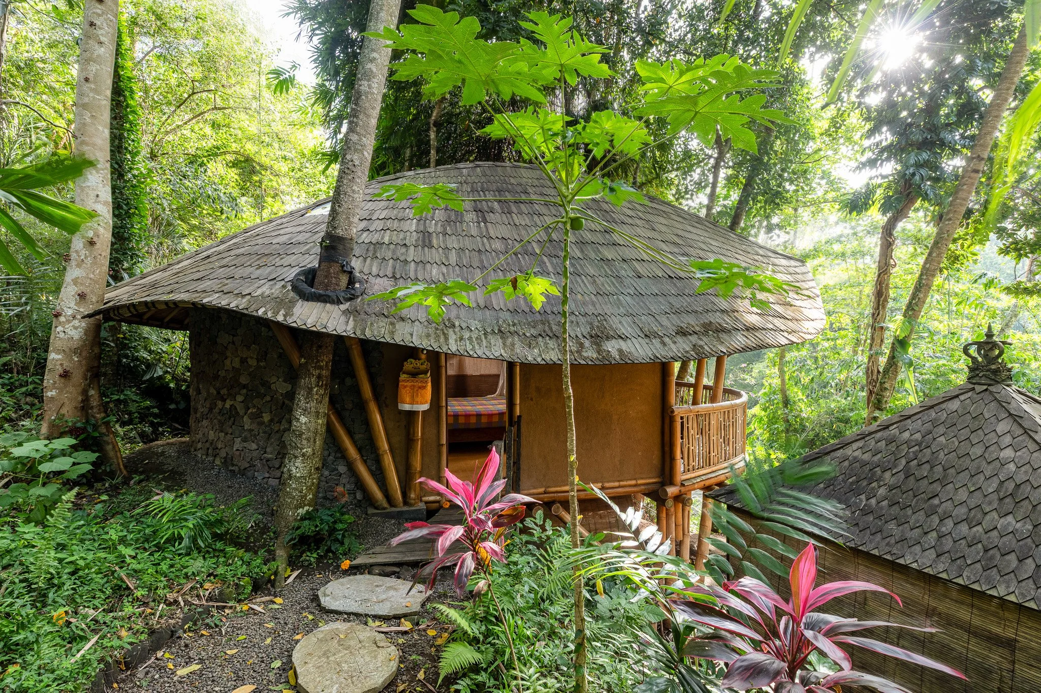 A circular treehouse built with bamboo and wood, situated in a lush green forest with dense trees and plants surrounding it. The treehouse has a thatched roof and a small balcony with a wooden railing.