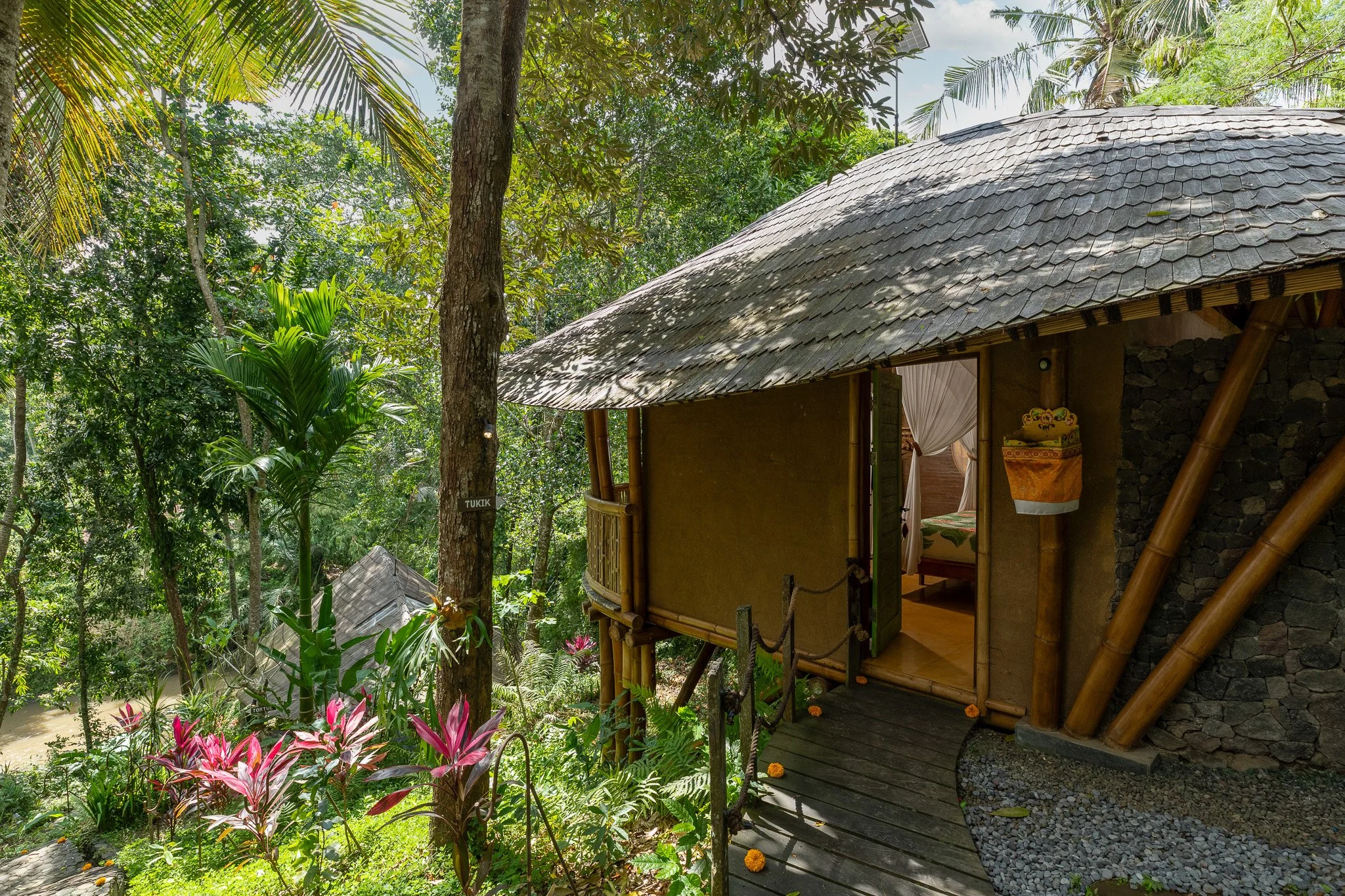 A treehouse with bamboo accents in a lush green forest, with a small porch, open door revealing a bed inside, and colorful plants around.