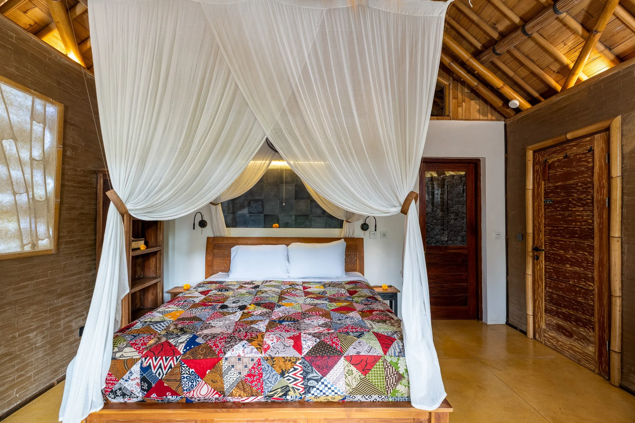 A bedroom with a four-poster bed draped with white canopy curtains, a patchwork quilt, wooden doors, and a textured wall behind the bed.
