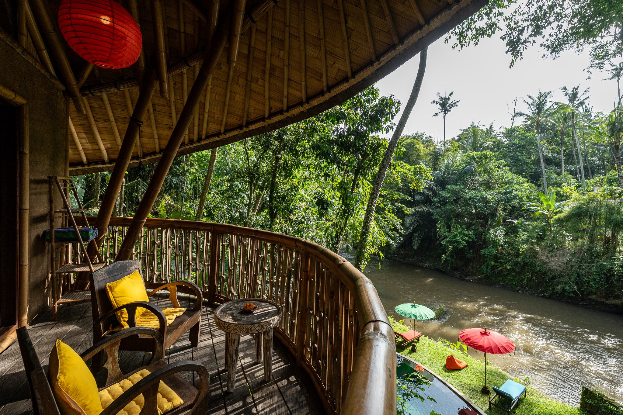Wooden balcony with yellow cushions, overlooking a river with colorful umbrellas and green outdoor chairs, surrounded by lush tropical trees.