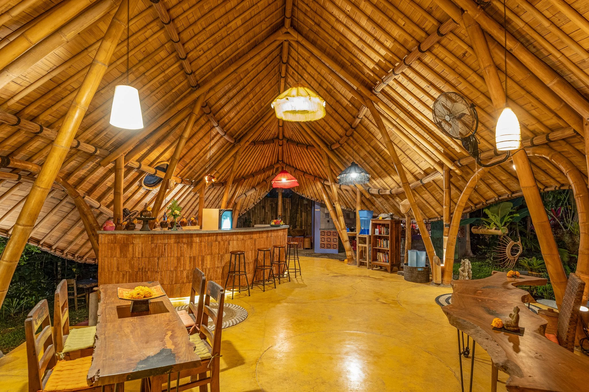Interior of a hut with bamboo roof, wooden furniture, colorful hanging lamps, a bar area, and tropical plants outside.