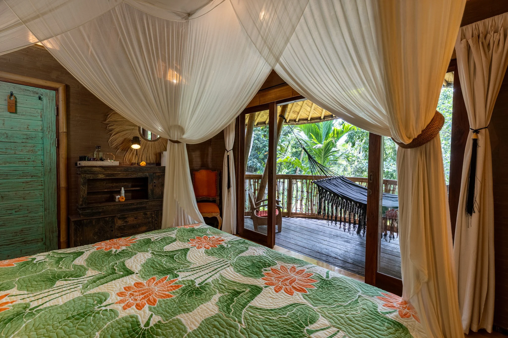 A tropical bedroom with a canopy bed, green and orange floral quilt, wooden furniture, and a balcony with a hammock and lush greenery outside.