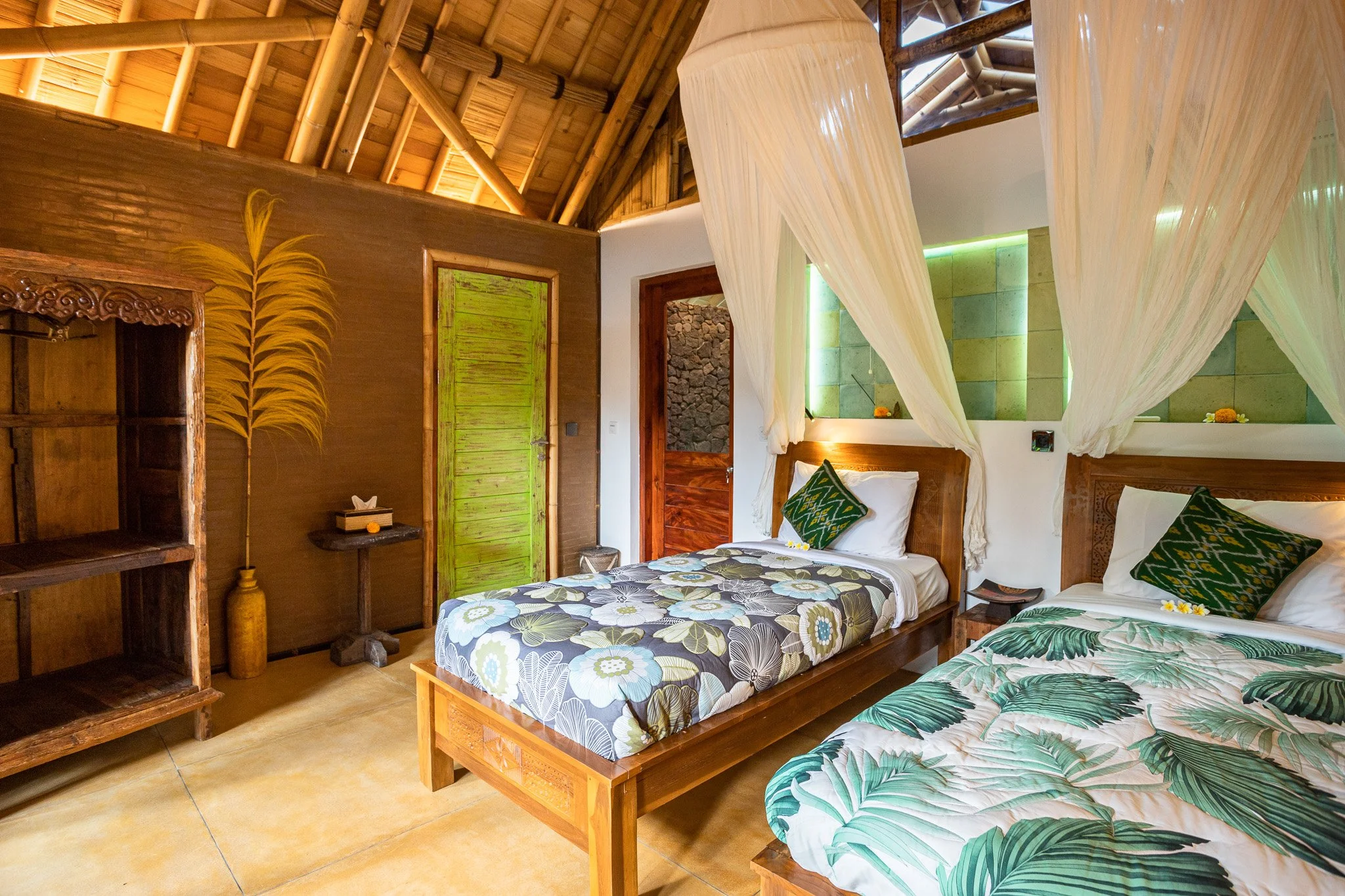 A bedroom with two beds featuring tropical leaf patterned bedding, mosquito netting canopy over each bed, wooden furniture, and a decorative wall with a fern motif. Tropical decor with warm lighting and natural wood accents.