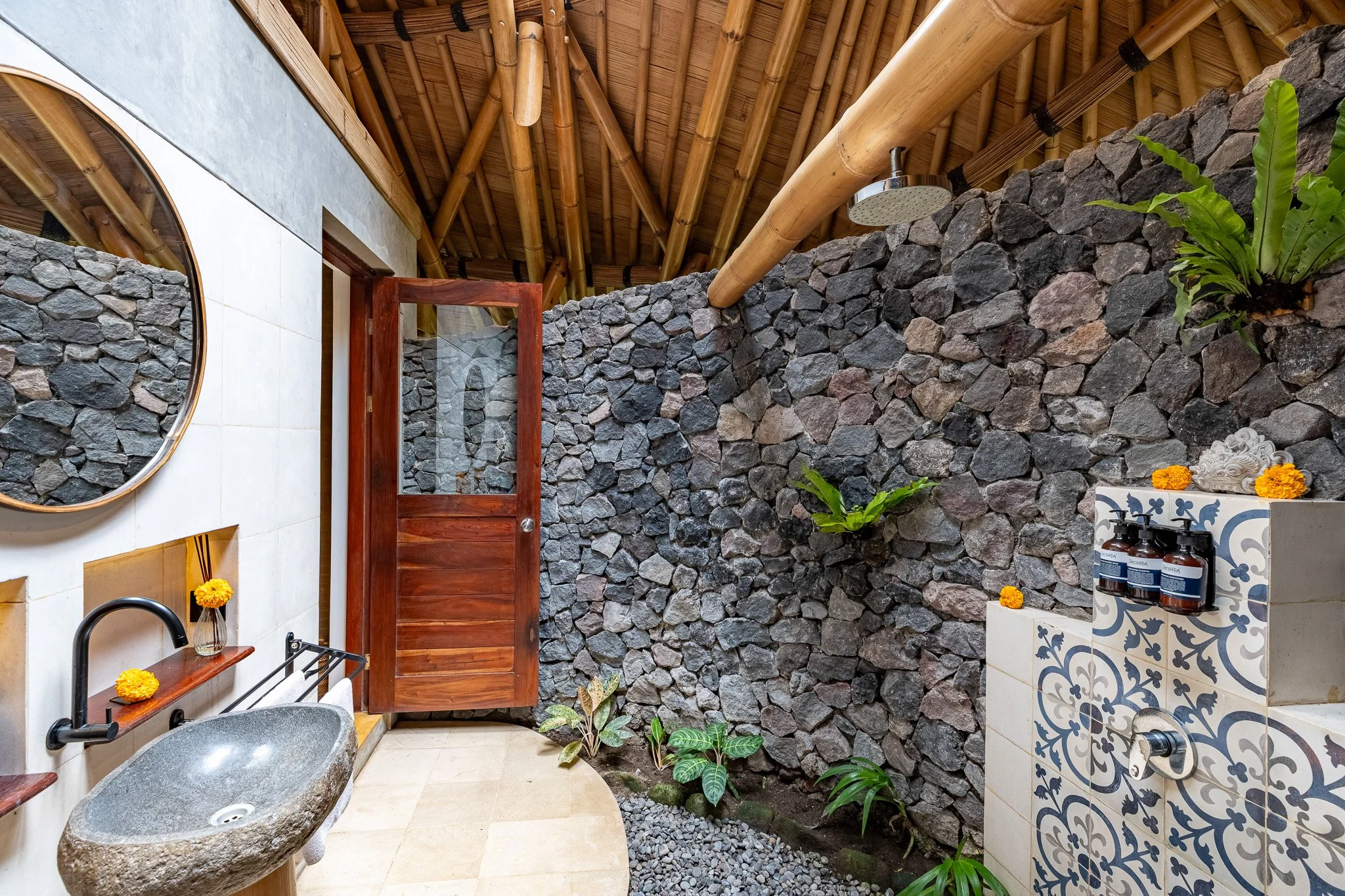 Interior of a bathroom with a stone wall, wooden ceiling with bamboo beams, a round mirror, a stone sink with a black faucet, a small shelf with yellow flowers, and a shower area with bottles of soap or shampoo and decorative flowers.