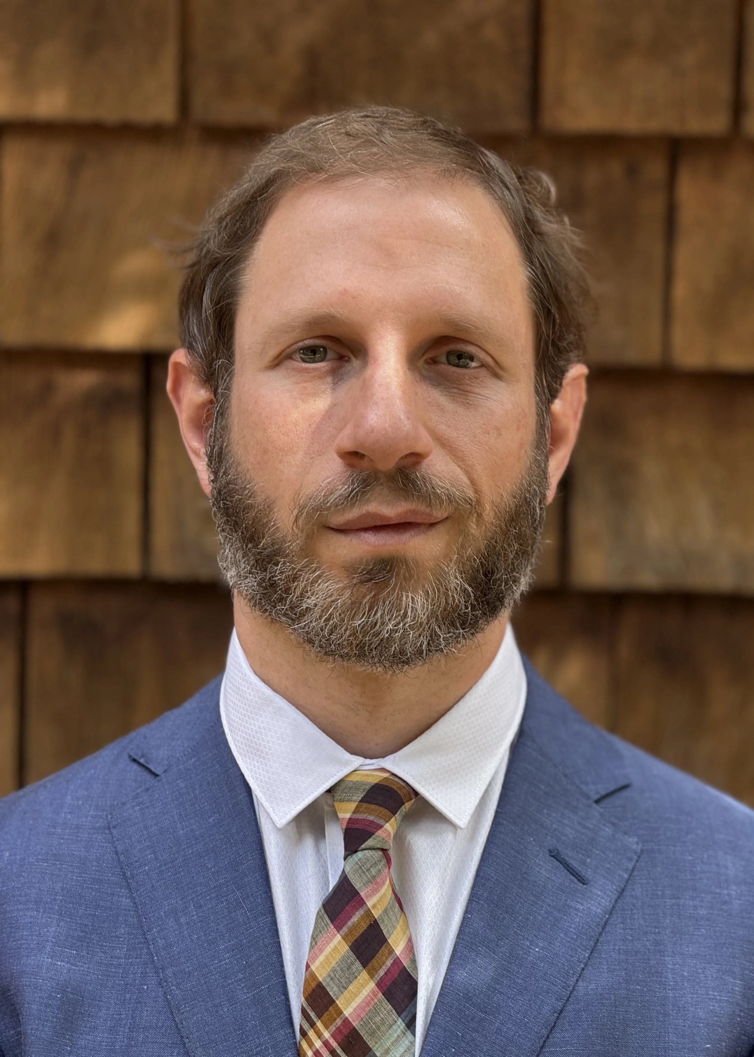Matthew J. Silberman, a Jewish man with a beard in a blue suit, white shirt, and a plaid tie standing in front of a wooden wall in Los Angeles, CA.
