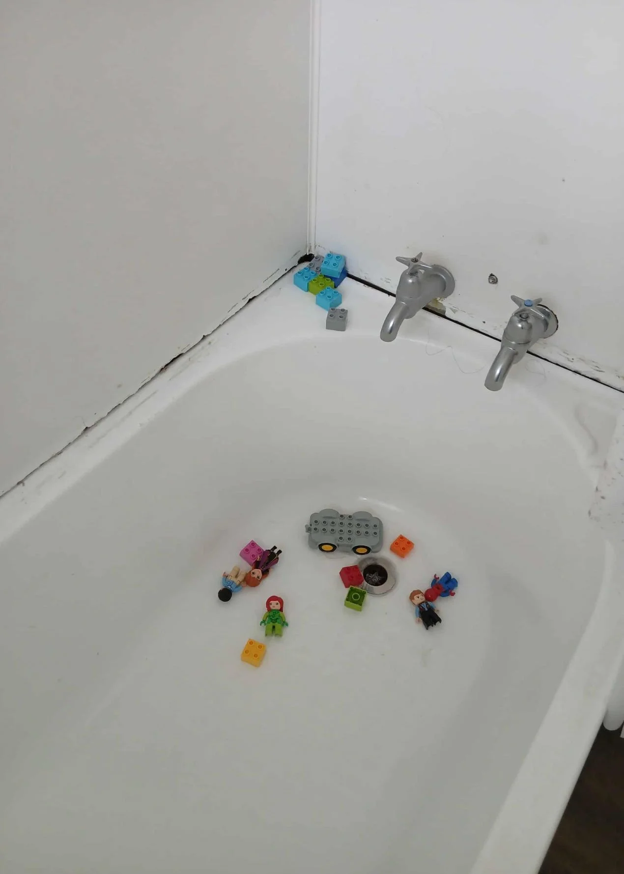 Messy bathtub with scattered LEGO toys, including mini-figures, blocks, and a small vehicle, with two silver faucet fixtures on the tiled wall.