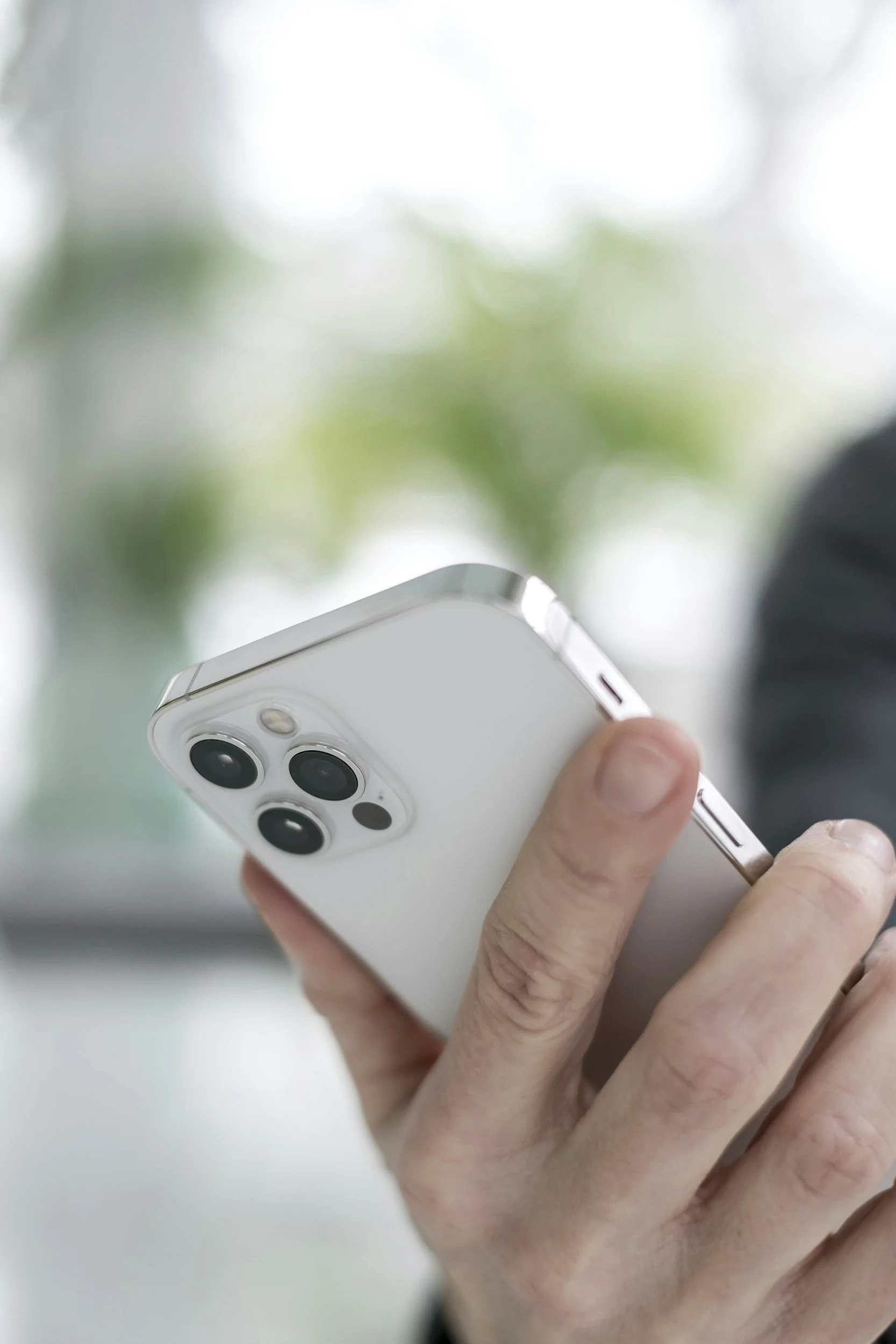 Person holding a white smartphone with three camera lenses on the back, in an indoor setting with blurred green plants in the background.