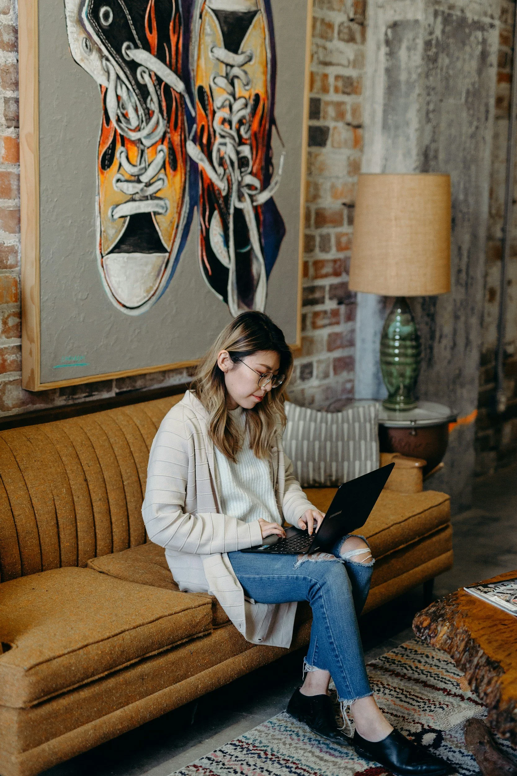 A woman with glasses, blonde hair, and casual clothes sitting on a vintage orange sofa working on a black laptop in a cozy, rustic room with exposed brick walls, a large colorful sneaker painting, a green ceramic table lamp, and a patterned rug.